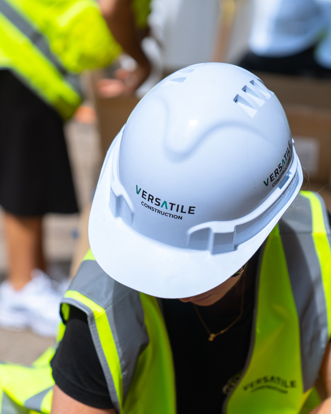 Close-up of a white construction helmet with the logo 'Versatile Construction' being worn by a person in a neon yellow safety vest at a construction site, with blurred background and other workers.