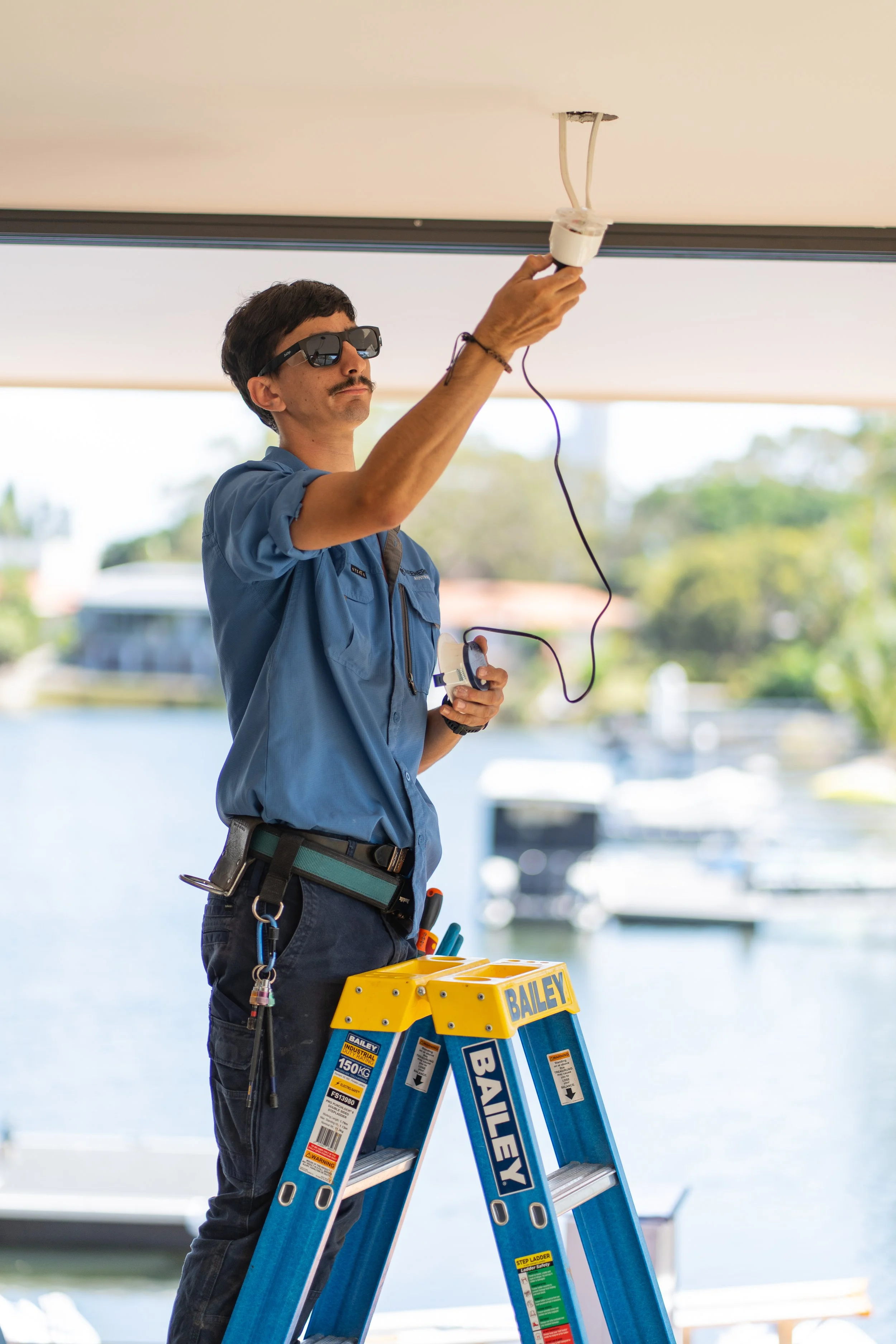 A man wearing sunglasses and a blue shirt standing on a ladder, working on a ceiling light fixture near a window with a view of water and boats outside.