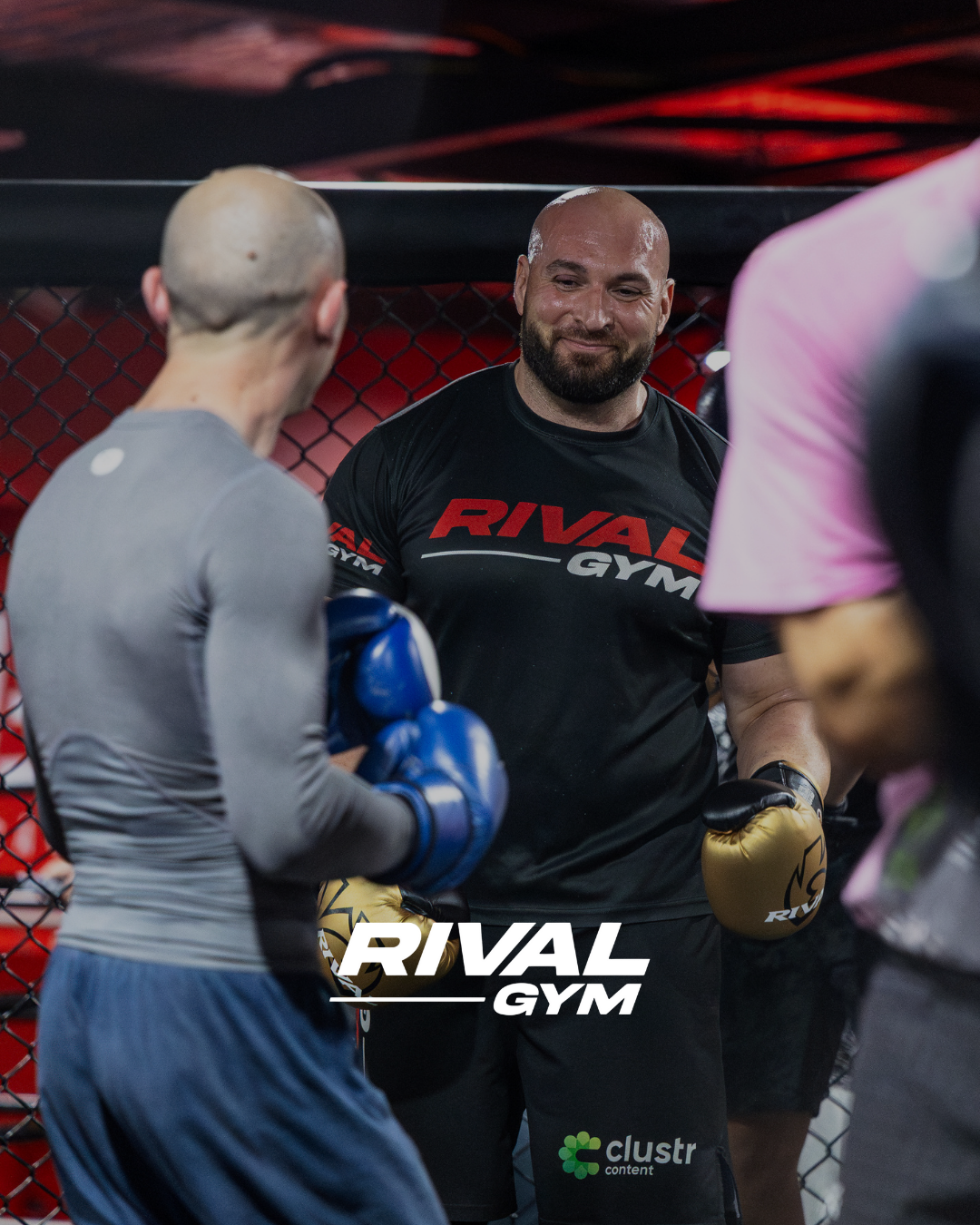 A group of MMA fighters in a training session at Rival Gym, with a man in black gloves smiling between two fighters, one with blue gloves and another with pink gloves, inside an octagon cage.