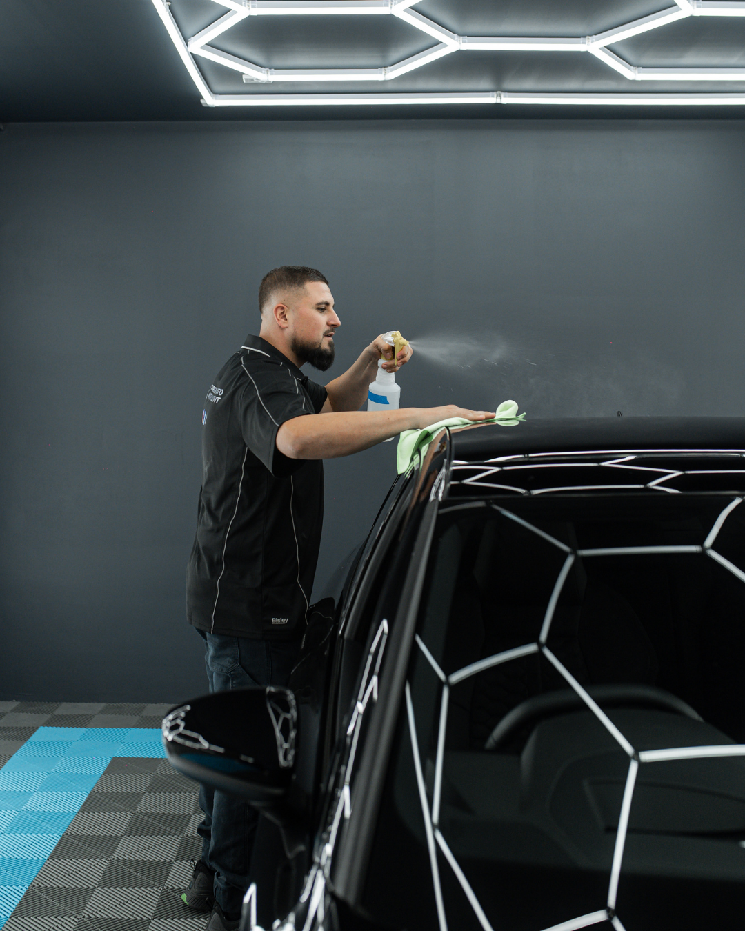 A man cleaning and detailing a black car with a white geometric pattern in an indoor garage or detailing shop, using a spray bottle and cloth.