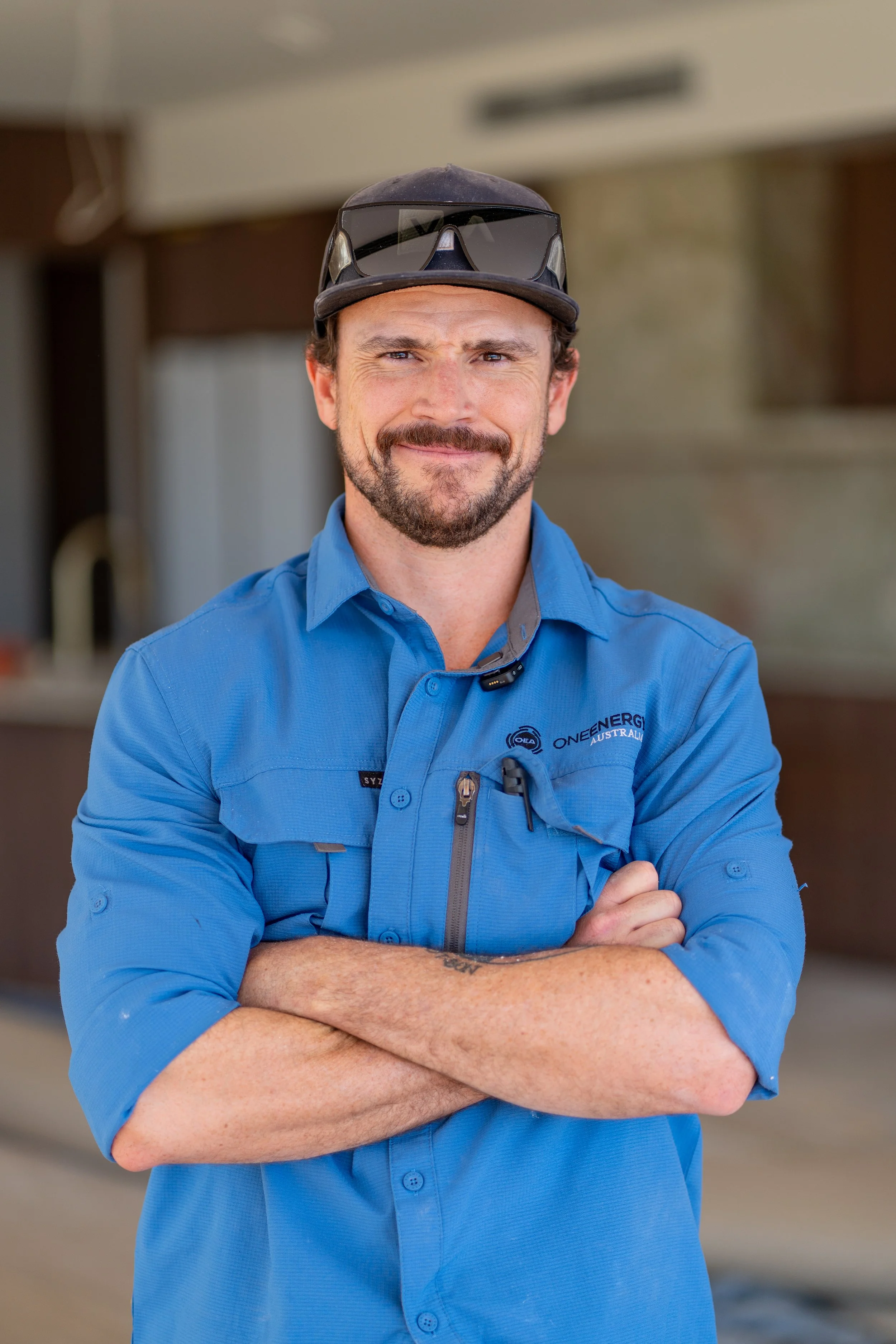 A man with a beard and mustache wearing a blue work shirt and a dark cap with sunglasses on top, standing with arms crossed in an indoor setting.