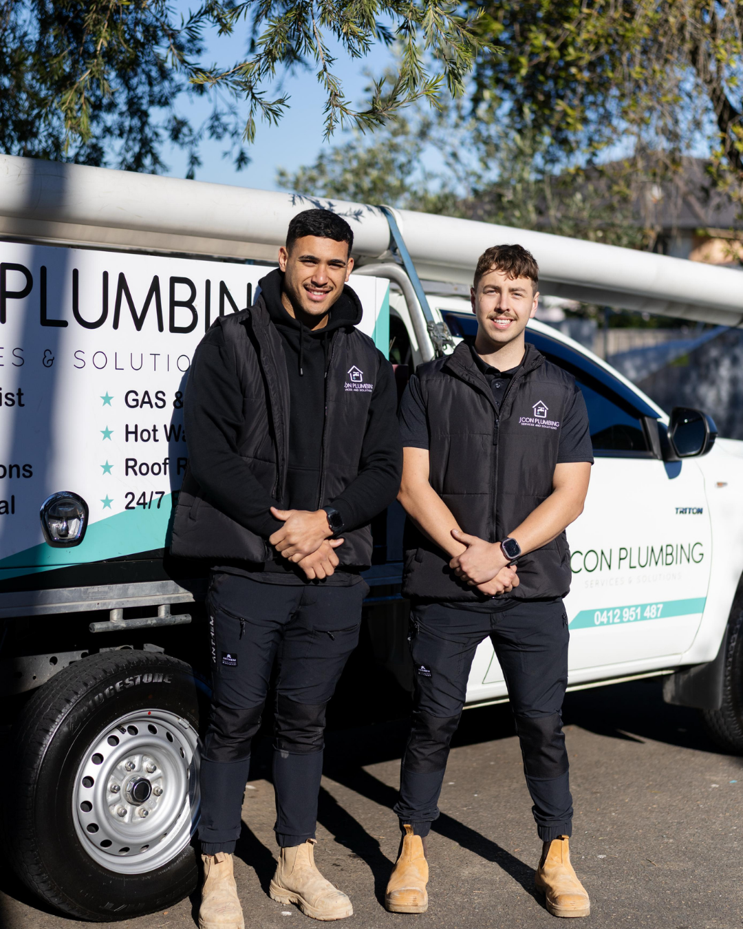 Two male plumbers standing in front of a plumbing service van, smiling at the camera, wearing work uniforms and tan work boots.