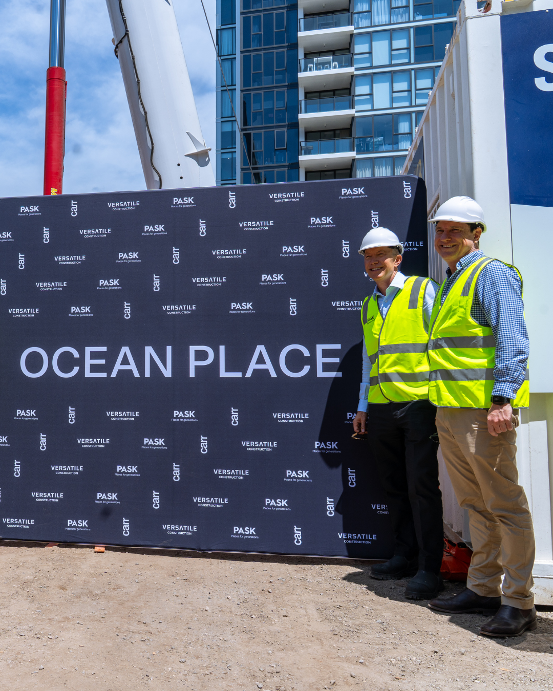 Two construction workers in yellow safety vests and white hard hats standing in front of a sign that reads "Ocean Place" at a construction site with a high-rise building in the background.