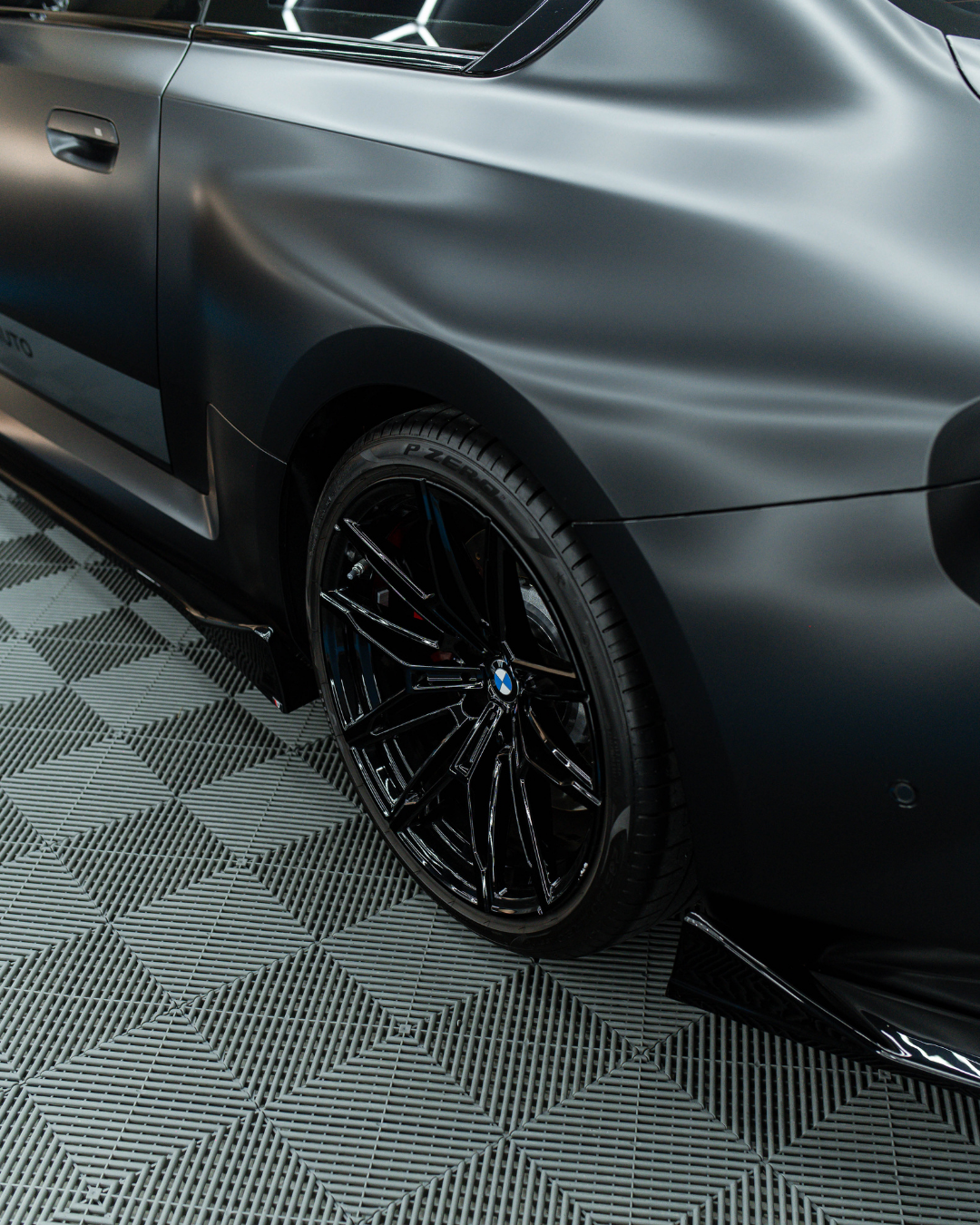 Close-up of a black BMW car tire and wheel with a matte gray body, parked on a textured black and gray floor.
