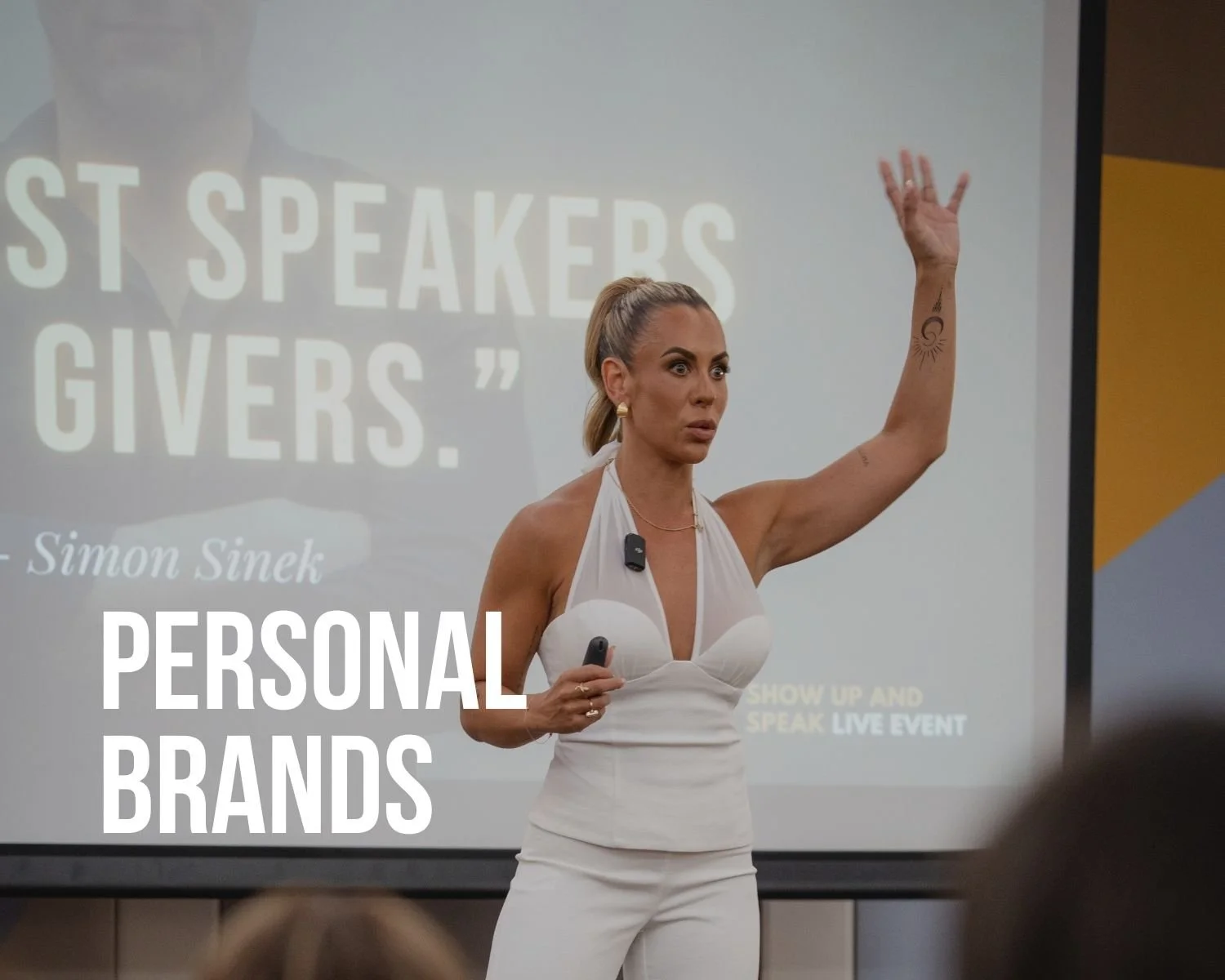 A woman with blonde hair in a ponytail giving a presentation in front of a screen with text about personal brands and speakers. She is wearing a white dress and holding a wireless microphone.