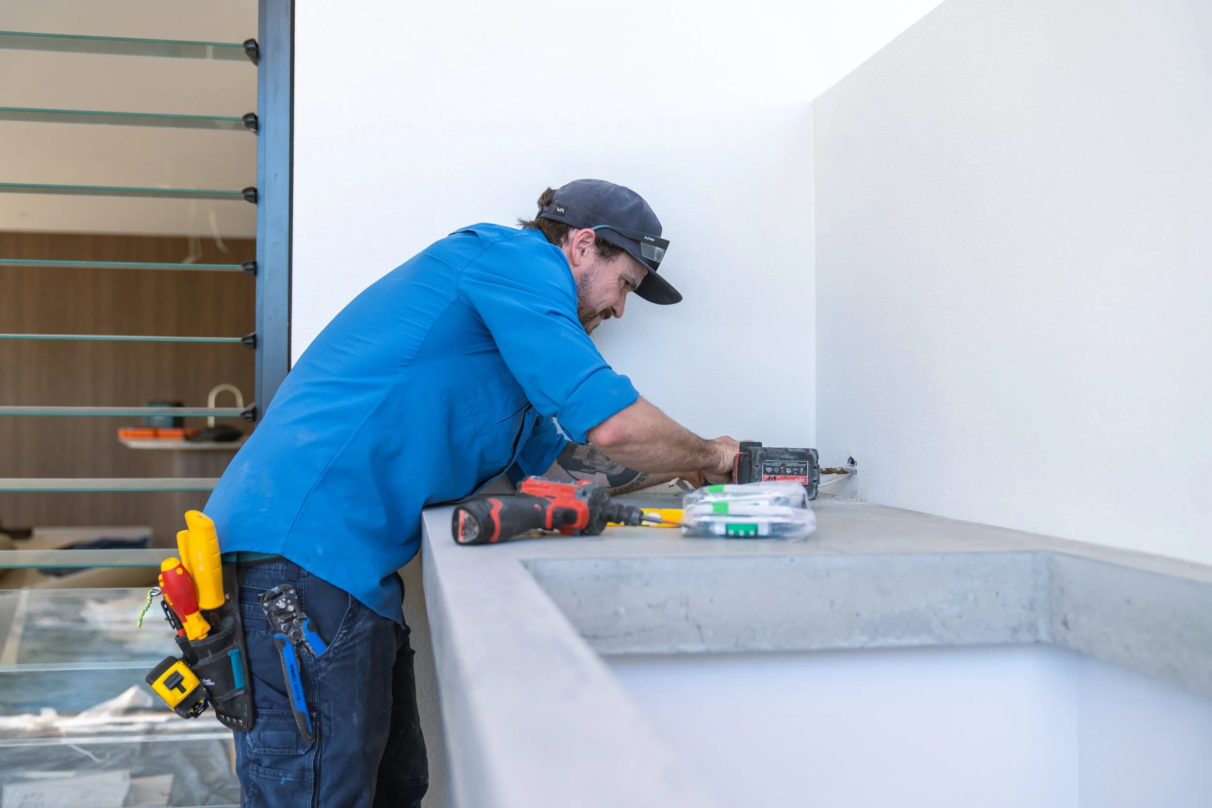 A man in a blue shirt and black cap installing or repairing something on a white wall, with tools on the concrete surface in front of him.