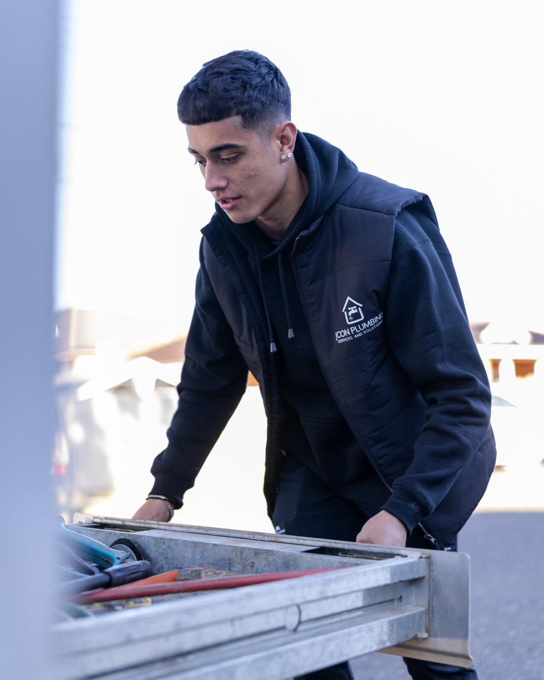 A young man in a black hoodie and vest working on plumbing outside during daytime.