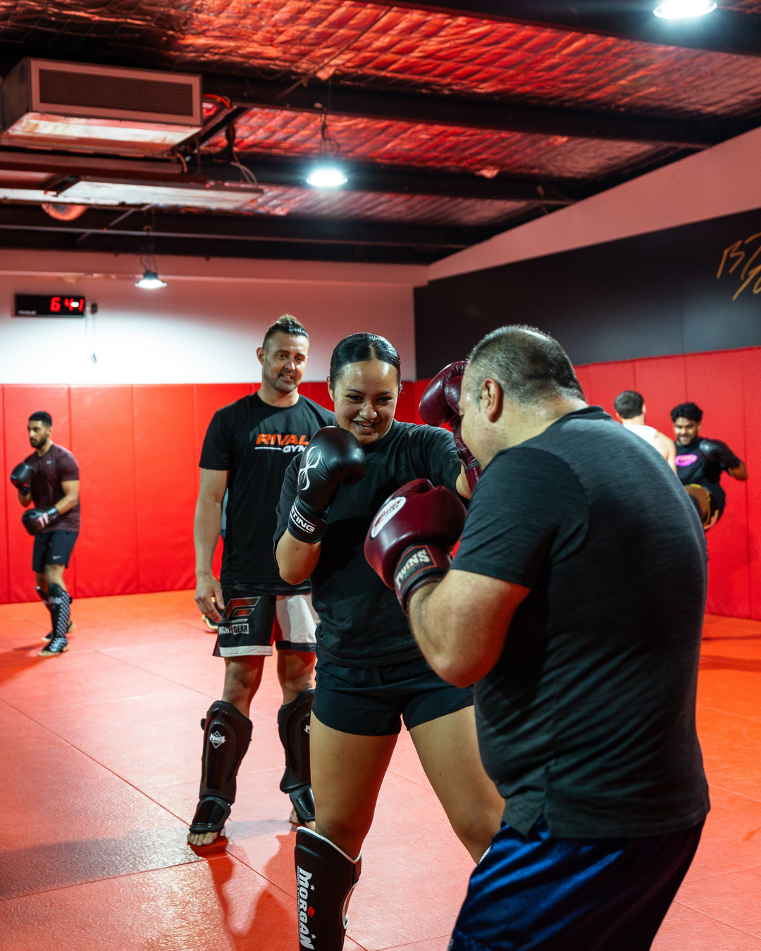 People practicing boxing and kickboxing in a gym with red padded walls, including a woman sparring with a man, and others training in background.