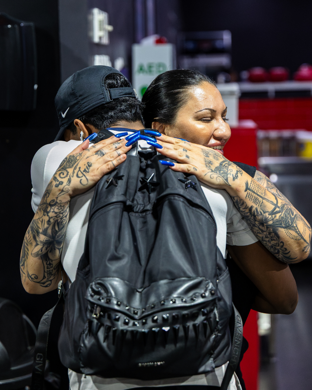 Two women hugging in a gym or locker room, one with extensive tattoos and blue acrylic nails, and the other in a black cap, carrying a black backpack with star-shaped embellishments.