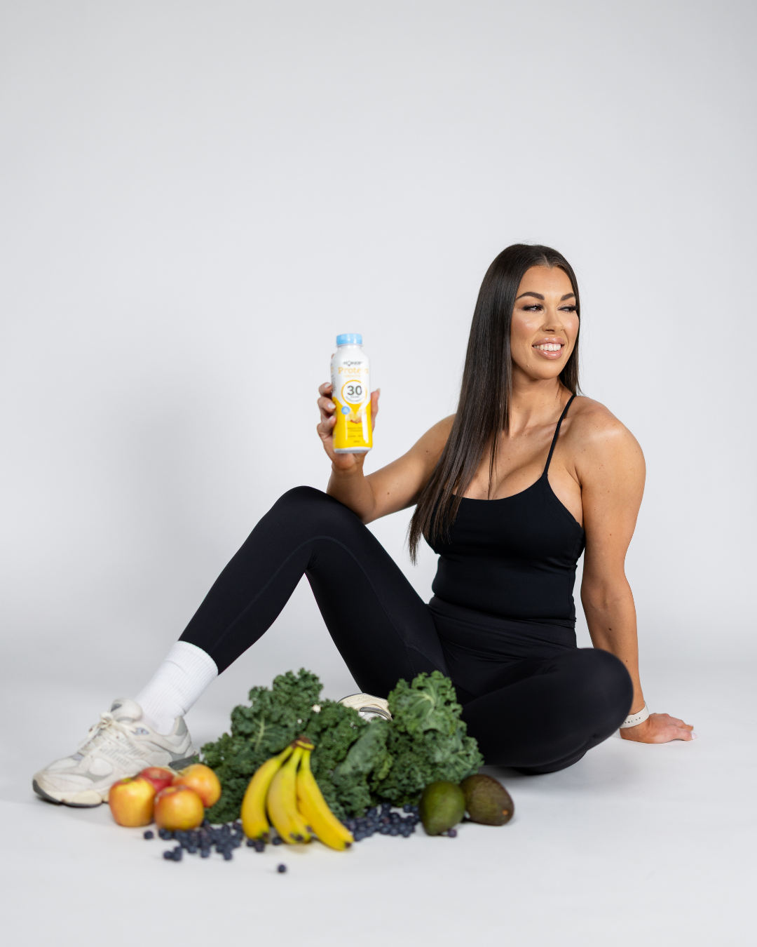 A woman in athletic clothing sitting on the floor surrounded by fresh fruits and vegetables, holding a bottle of sunscreen.