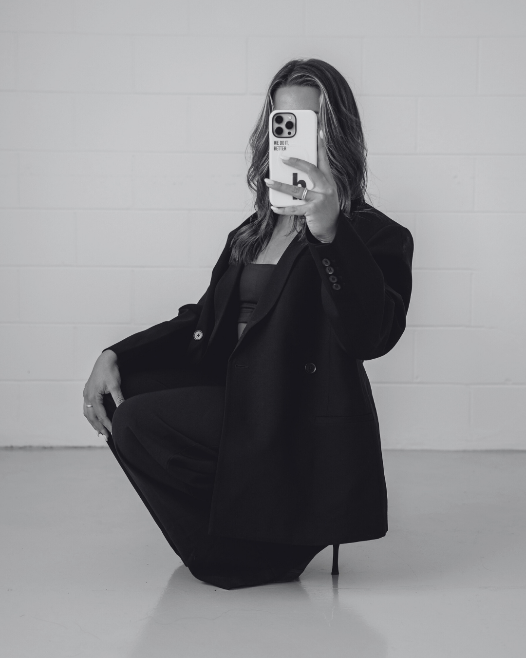 A woman with shoulder-length hair taking a mirror selfie with her phone, which has a case featuring text and a logo. She is dressed in a black blazer and black pants, with one knee raised and her hand resting on her knee. The background consists of a plain, light-colored wall.
