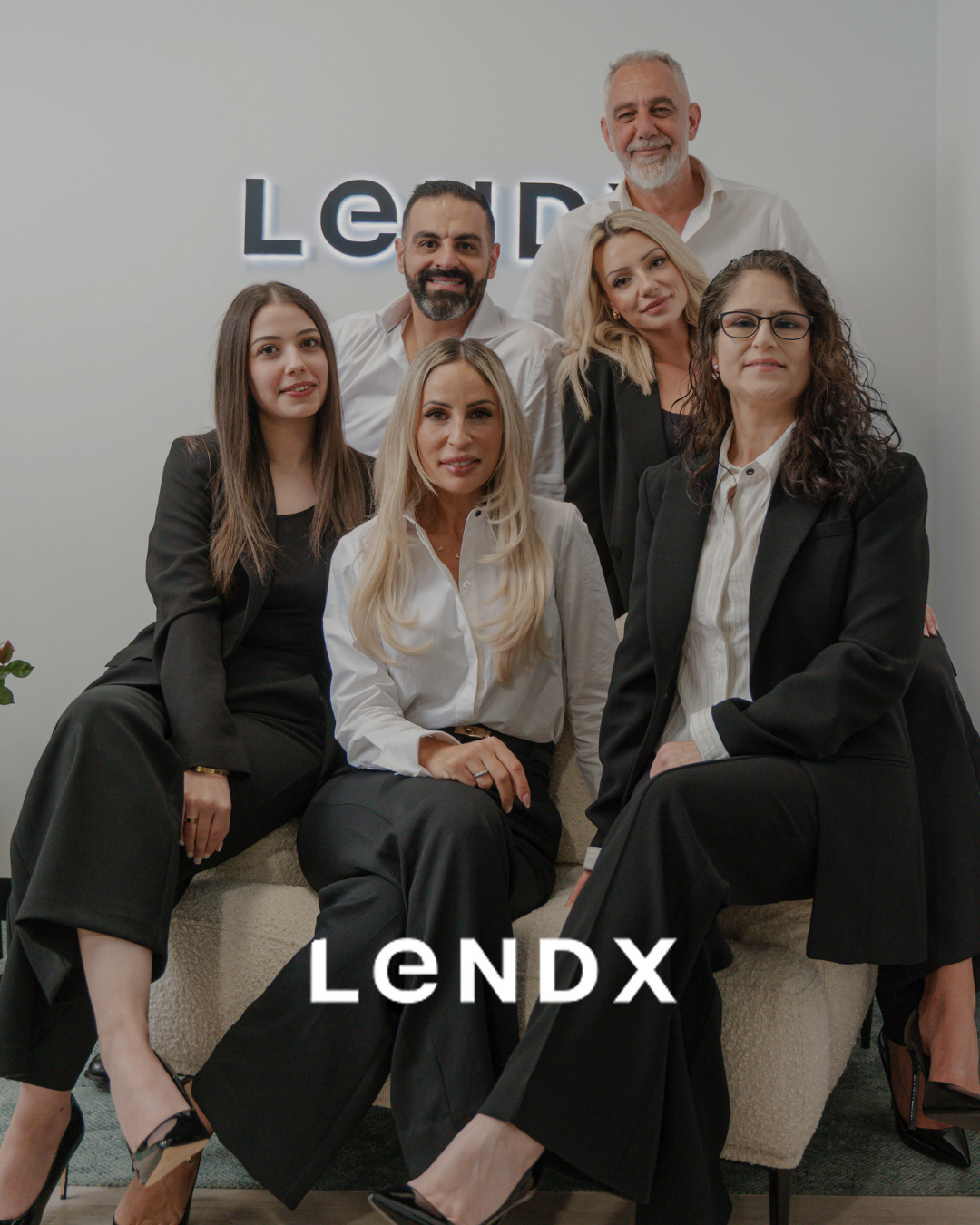 Group of six professionally dressed people in an office, posing for a photo with a sign reading "LENDX" in the background.