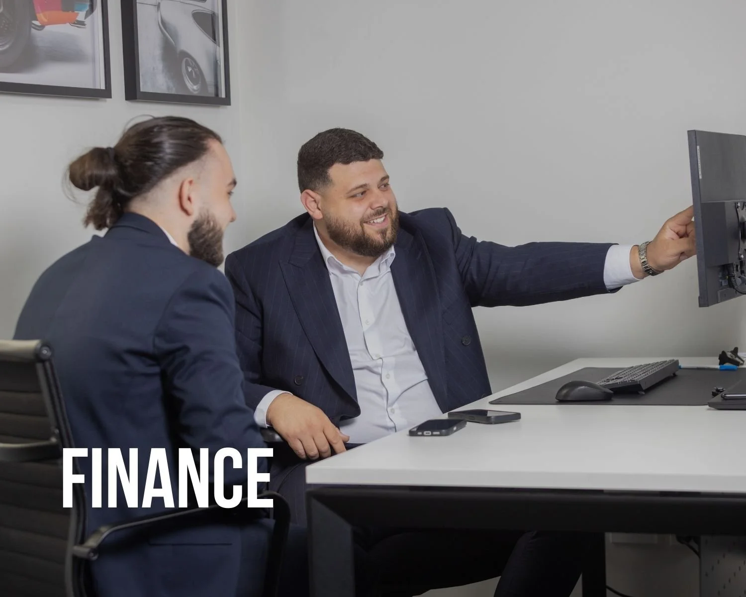 Two men in business suits are sitting at a desk in an office, looking at a computer screen and smiling. One man is pointing at the monitor. The word "FINANCE" is written at the bottom left corner of the image.