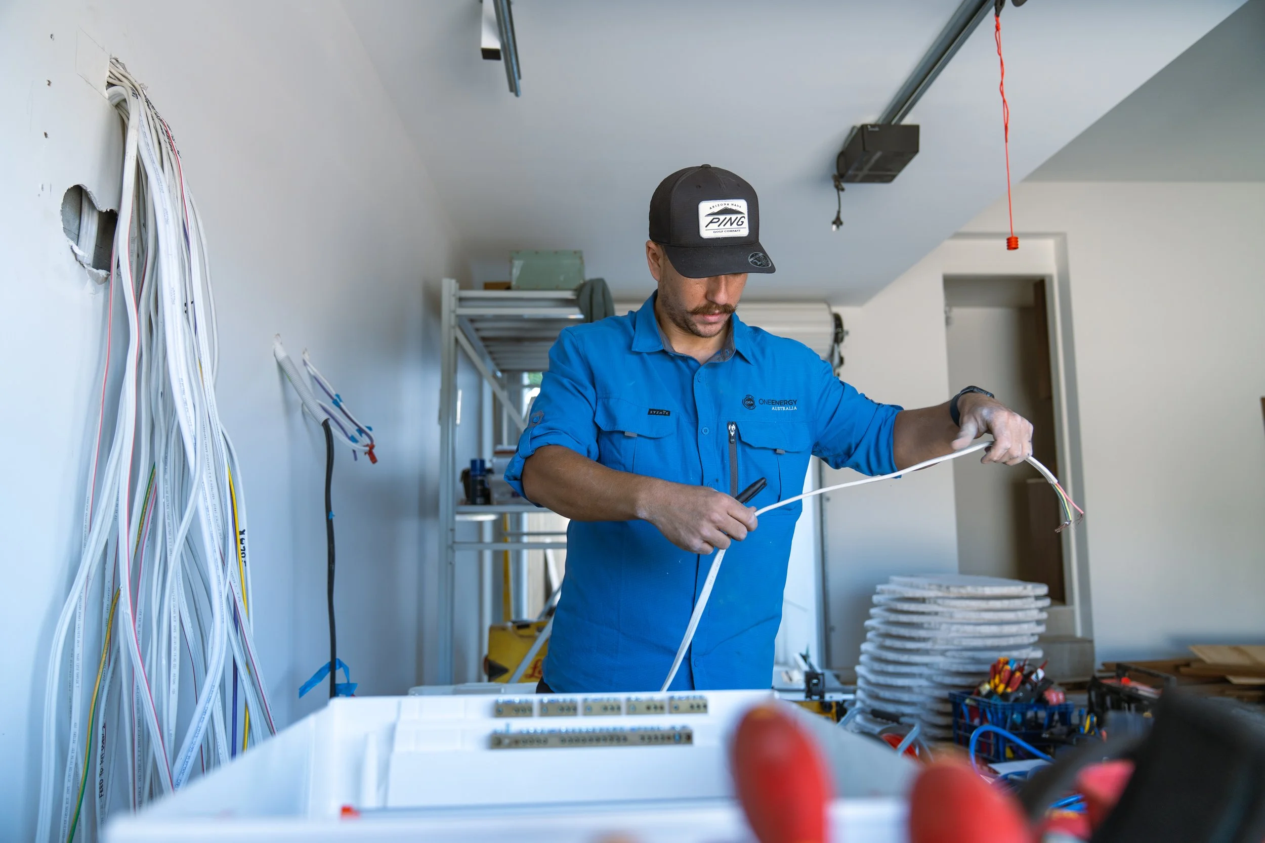 A technician in a blue work shirt handling electrical wires in a workshop with tools and cables around him.