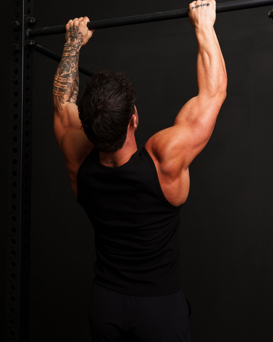 Man doing pull-ups on a black exercise bar, wearing a black tank top, with tattoos on his left arm, in a gym setting with a dark background.