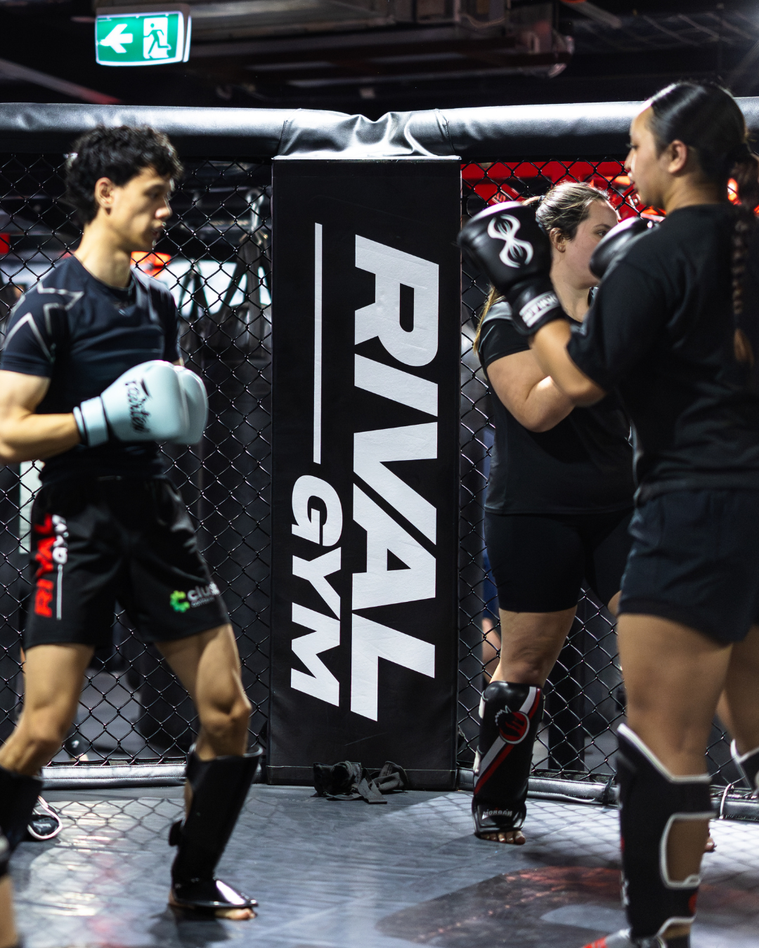 Women practicing mixed martial arts in a training cage, with one woman boxing and another woman receiving instruction.