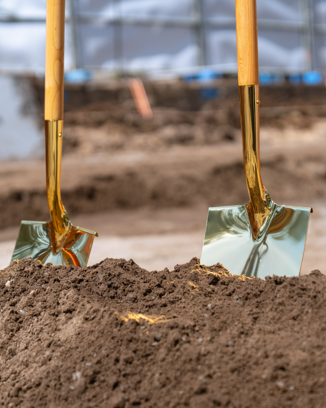 Two gold-colored shovels with wooden handles stuck in a mound of dirt at a construction site.