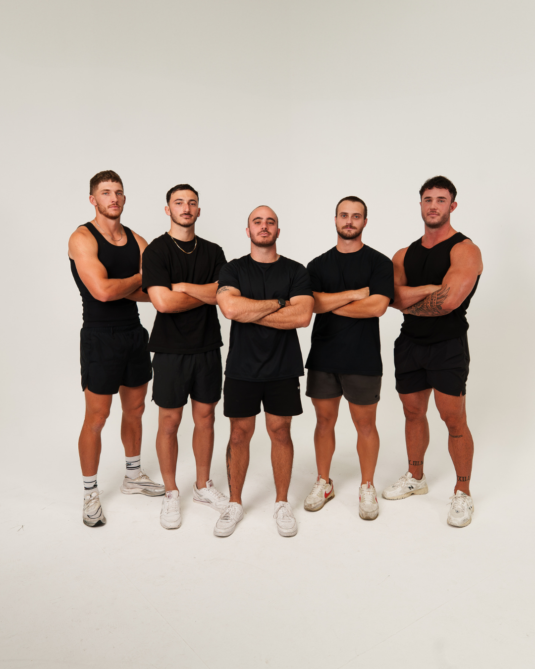 Five young men standing with arms crossed, wearing black athletic shirts and shorts, in a studio with a plain white background.