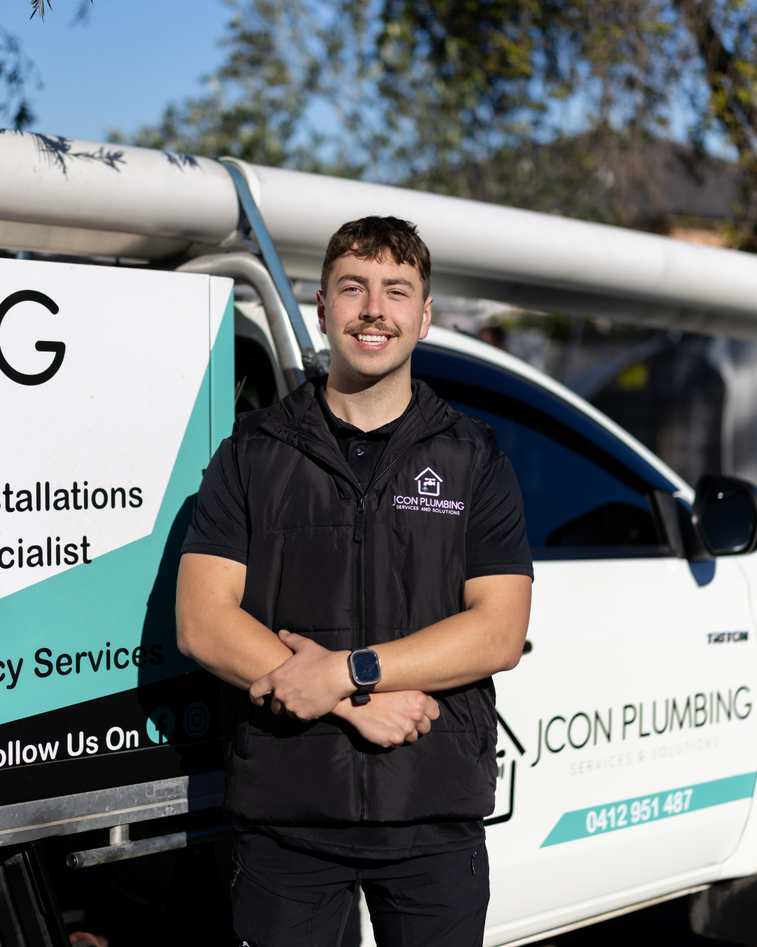 A smiling young man standing in front of a service truck for JCON Plumbing with plumbing equipment on top, wearing a black vest with the company logo.