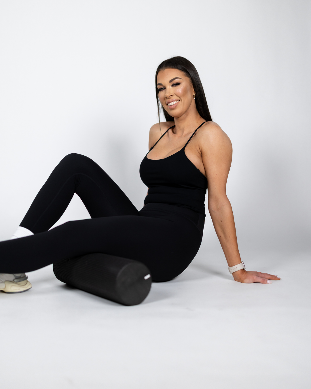 Woman in black workout clothes sitting on the floor with a black foam roller under her knee, smiling and winking at the camera.