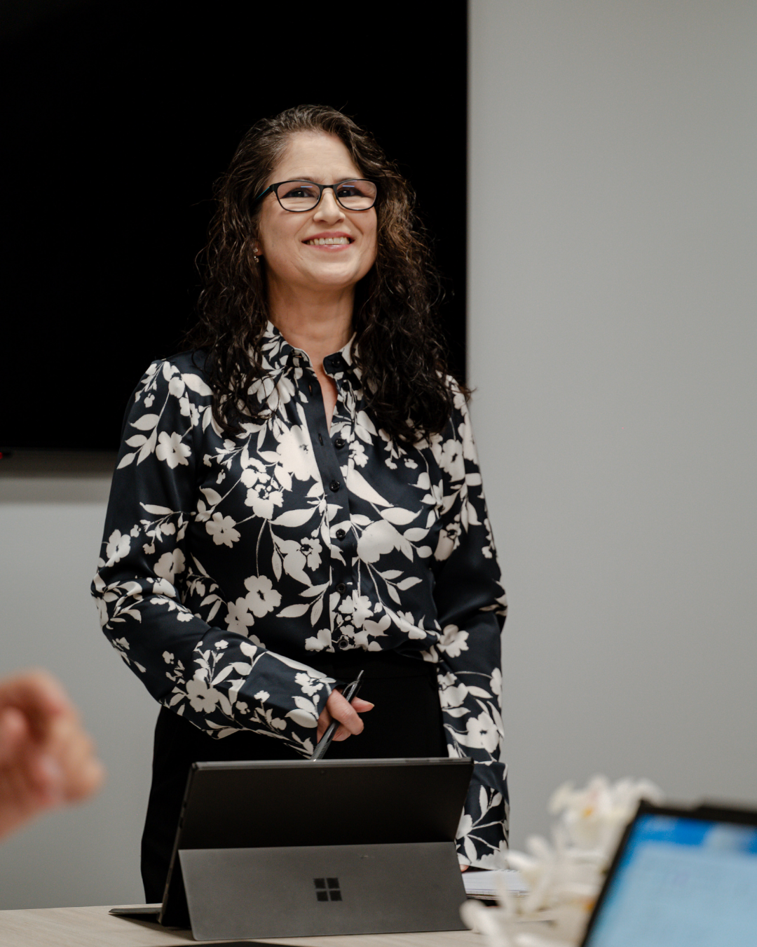 A woman with curly hair and glasses, wearing a black and white floral blouse, stands in a meeting room smiling.
