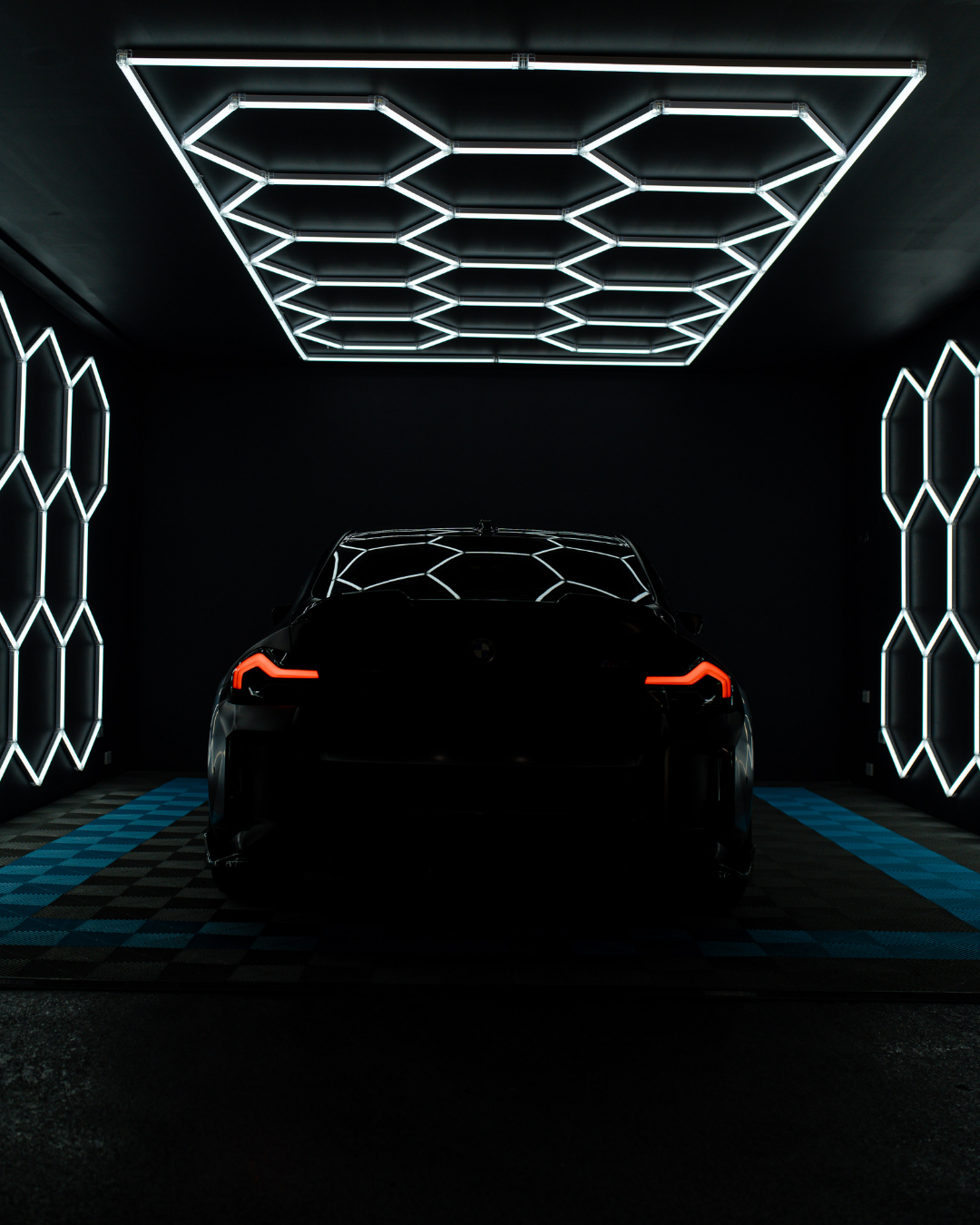 A dark room with black walls and a checkered floor, illuminated by geometric white neon light fixtures on the ceiling and walls, with the silhouette of a black car with illuminated taillights in the center.
