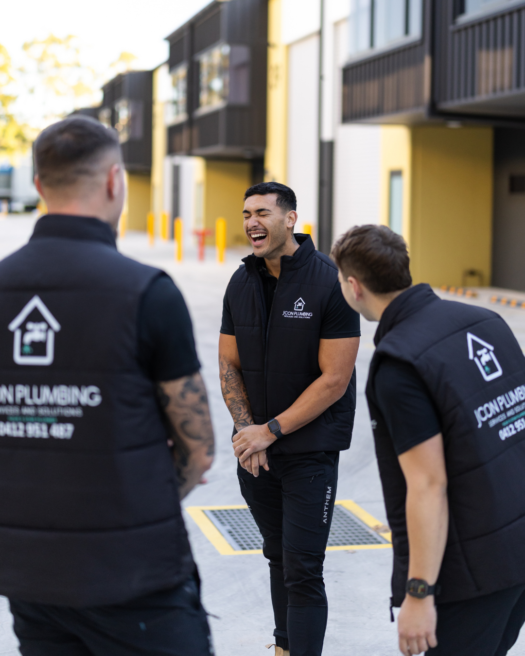 Three men in black uniforms are standing outside, with two of them facing away and one facing front, smiling and talking. They are in front of a building with black and yellow exterior walls.
