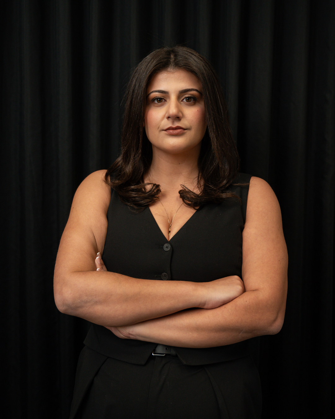 Confident woman with dark hair and a black sleeveless top standing with arms crossed against a black curtain backdrop.