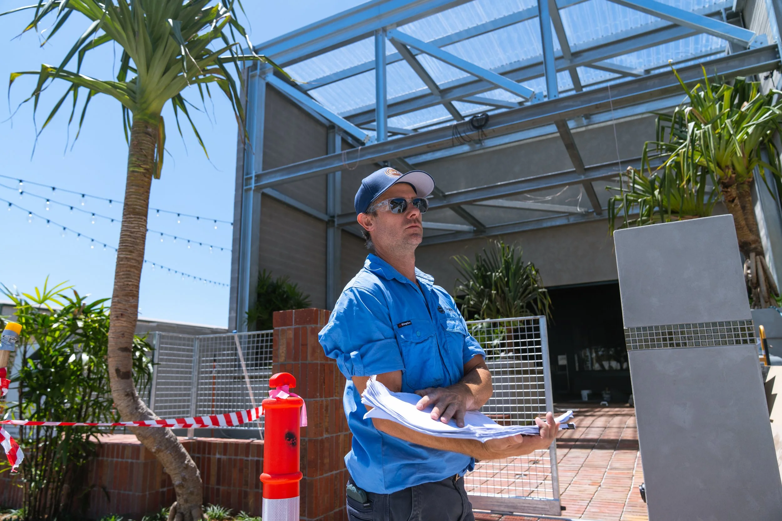 A man wearing sunglasses, a blue shirt, and a baseball cap standing outdoors near construction or renovation site, holding papers and looking serious.