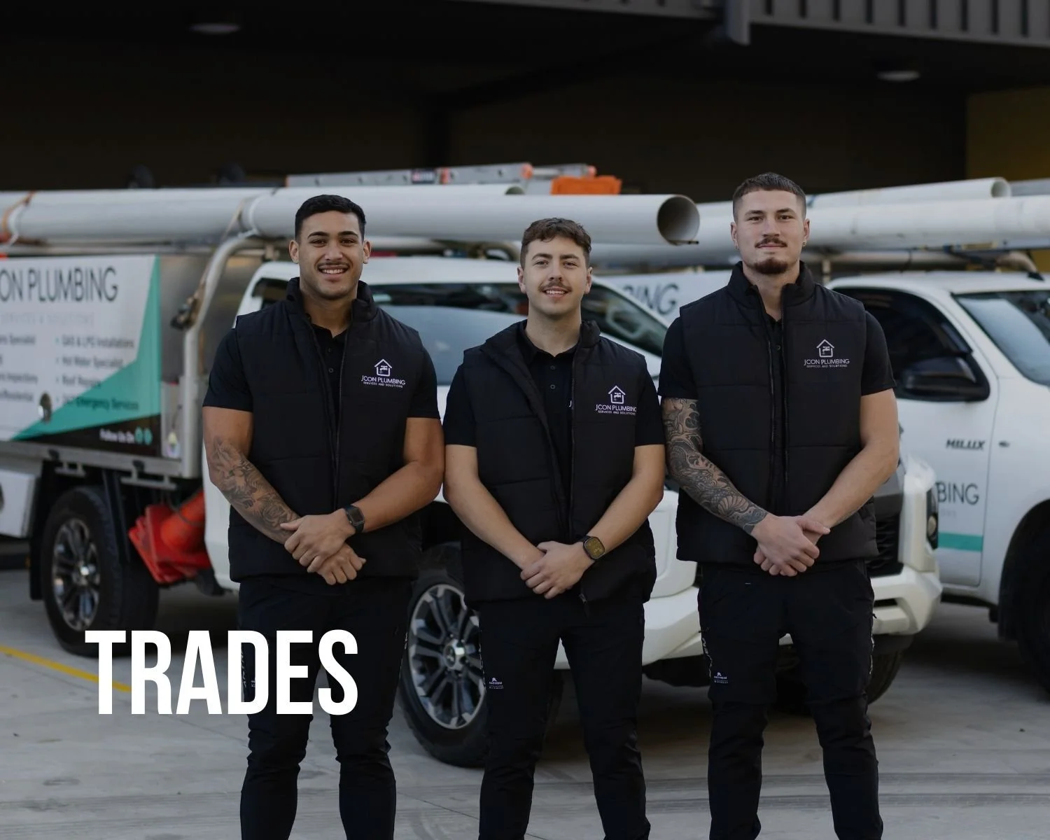 Three male workers in black uniforms standing in front of a company branded truck with pipes on top. They are smiling and standing with their hands clasped in front of them. The word 'TRADES' is written in white at the bottom left corner.