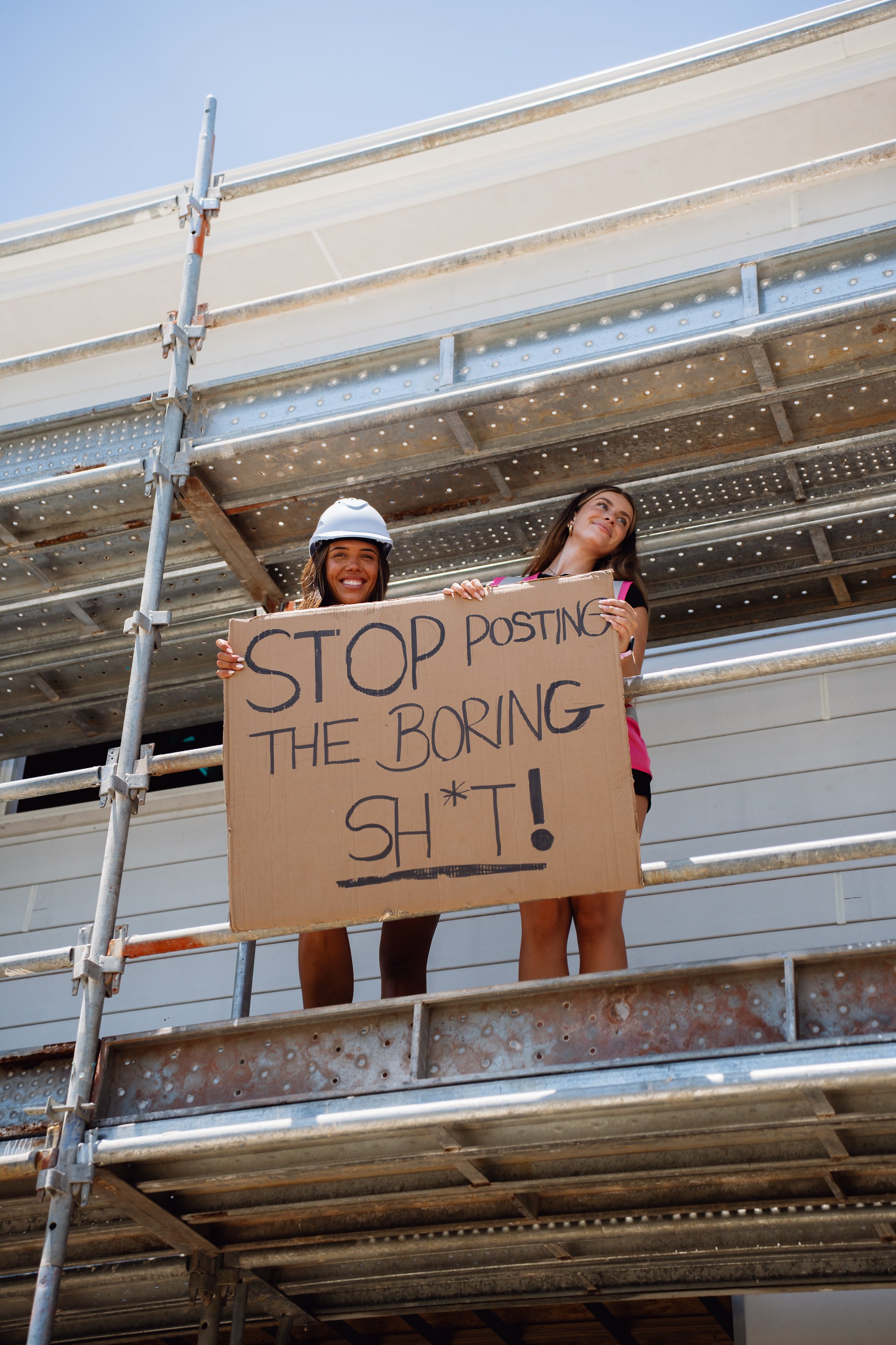 Two young women standing on construction scaffolding holding a cardboard sign that reads, 'Stop posting the boring sh*t!'
