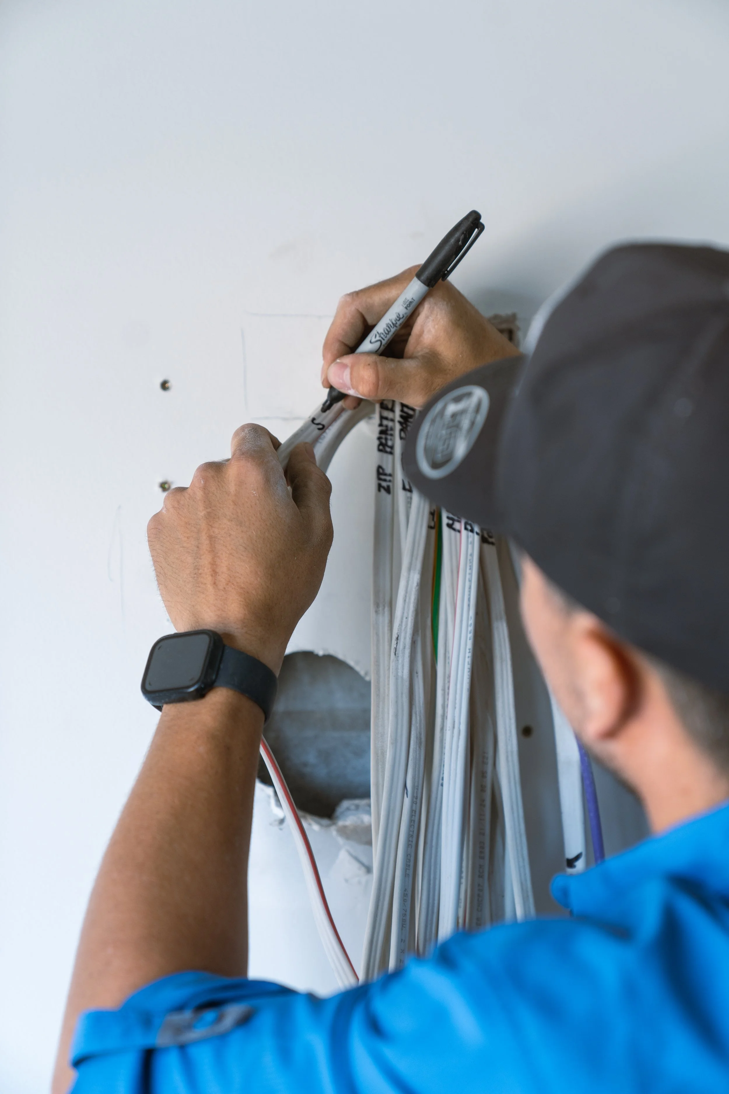 A person with a black cap and a blue shirt is writing on an electrical box with a black marker, surrounded by electrical wires.