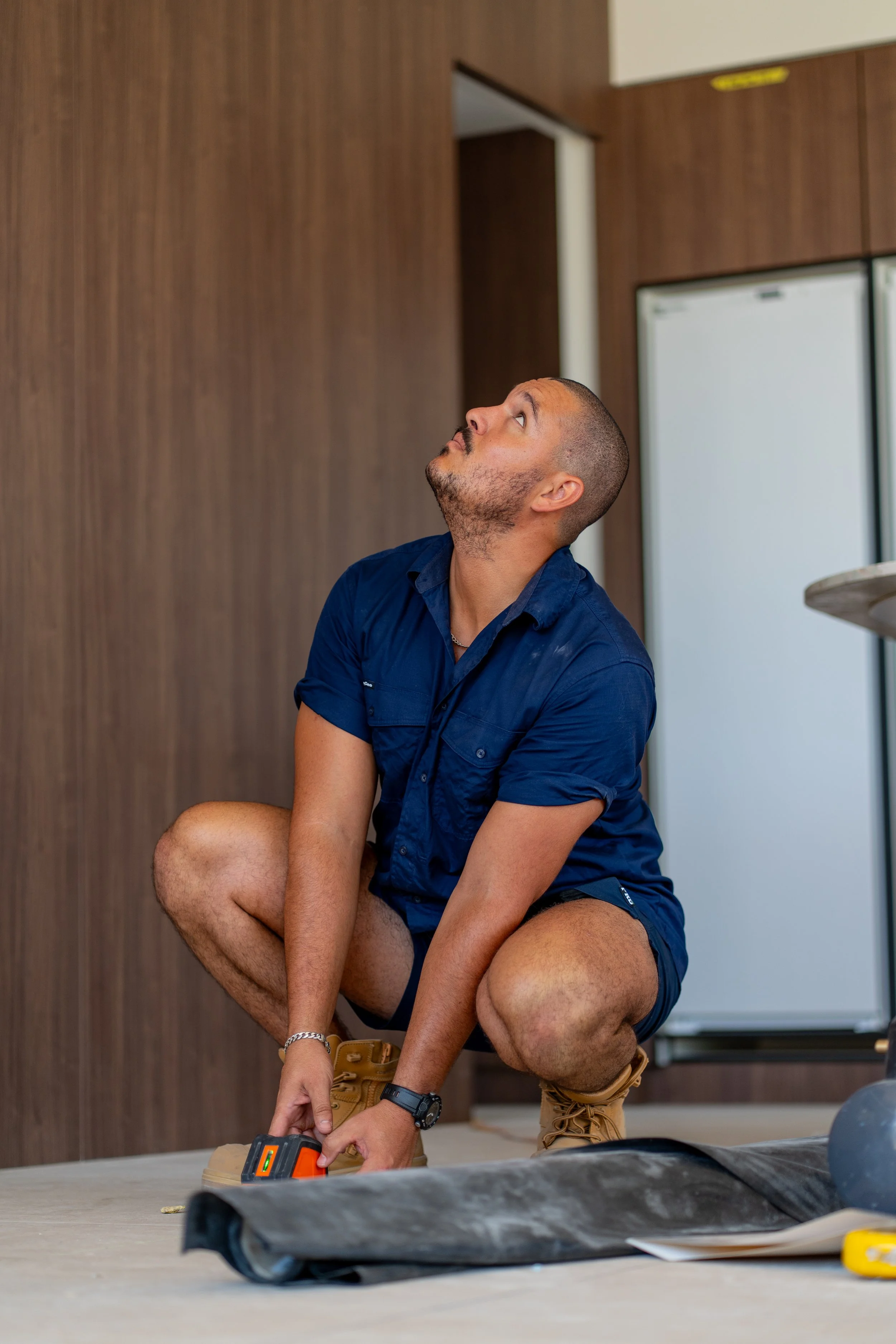 A man crouching on the floor, holding a measuring tape, in a room with wooden wall panels. He is looking upward, with a concentration on his face, wearing a navy blue shirt, shorts, boots, and a wristwatch.