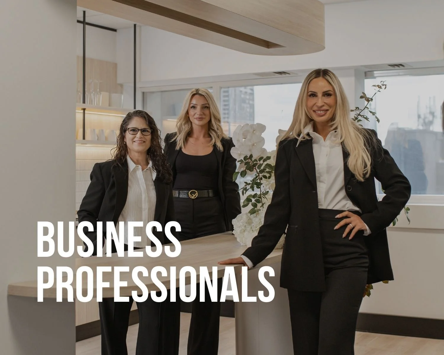 Three women in business attire standing in an office with a modern decor, large windows, and plants, smiling at the camera with the text 'Business Professionals' overlaid.