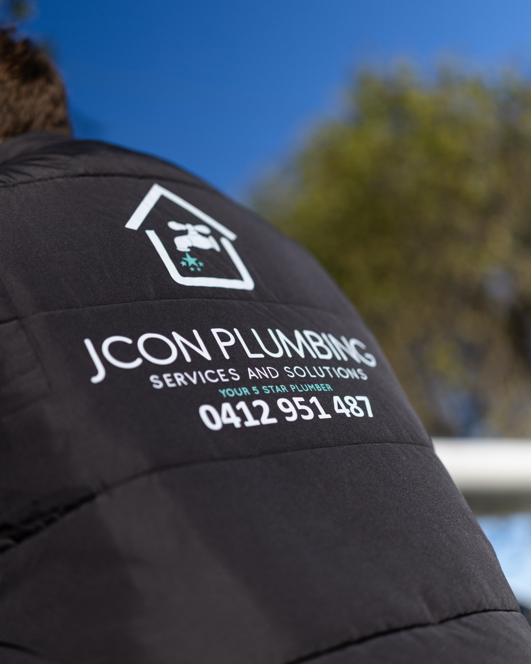 Close-up of a black jacket with a logo and contact information for JCON PLUMBING Services and Solutions, including phone number 0412 951 487. The background shows a blurred outdoor scene with blue sky and tree leaves.