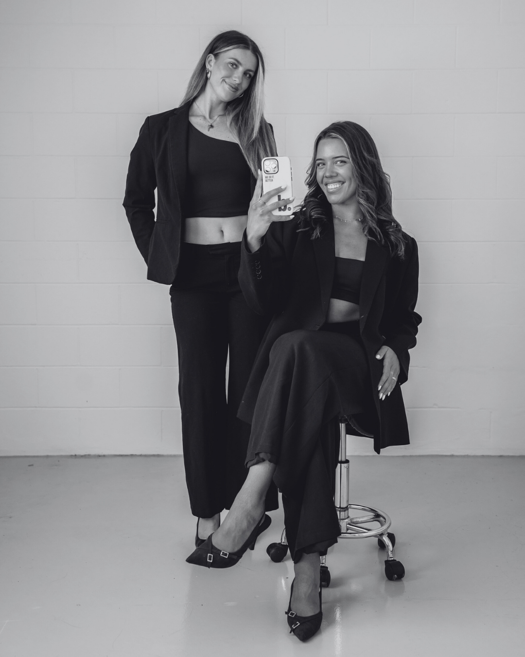 Two women in black outfits taking a selfie in front of a plain wall. One is standing and the other is sitting on a stool. Both are smiling and looking at the camera.