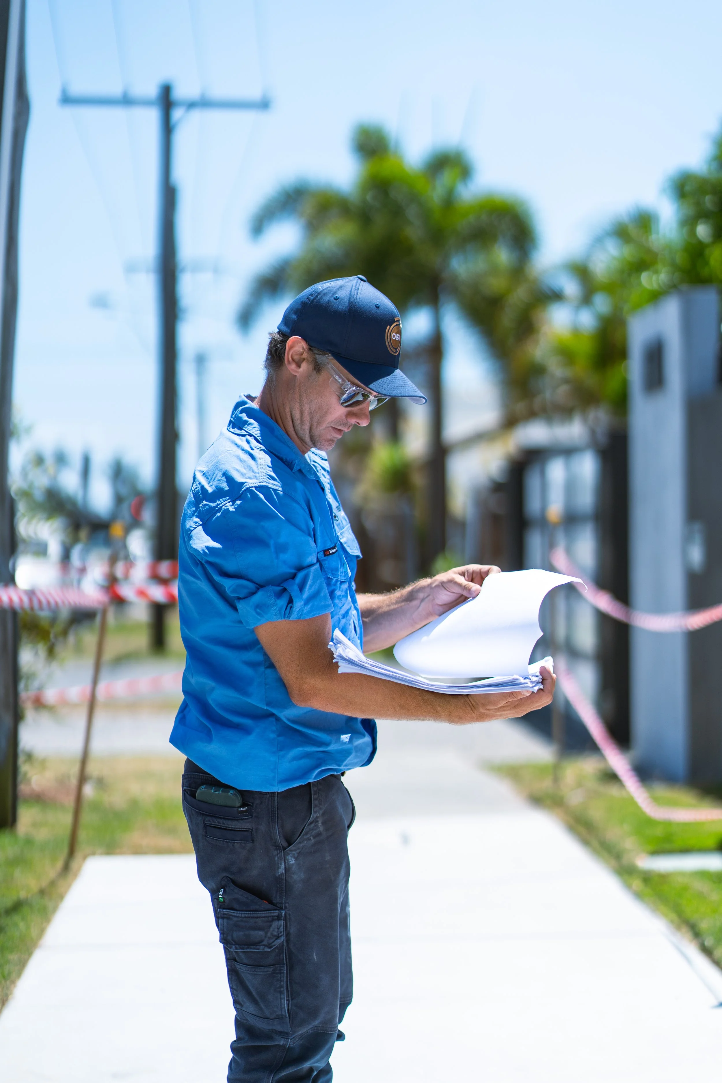 A man wearing a blue shirt, black pants, sunglasses, and a black cap is reading a stack of papers outdoors on a sunny day, with palm trees and utility poles in the background.