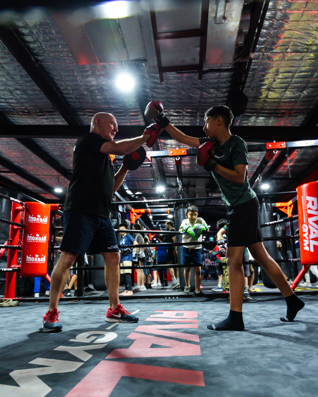 A boxing trainer and a young boxer spar in a boxing gym, with both wearing boxing gloves, while other boxers and trainers train in the background.