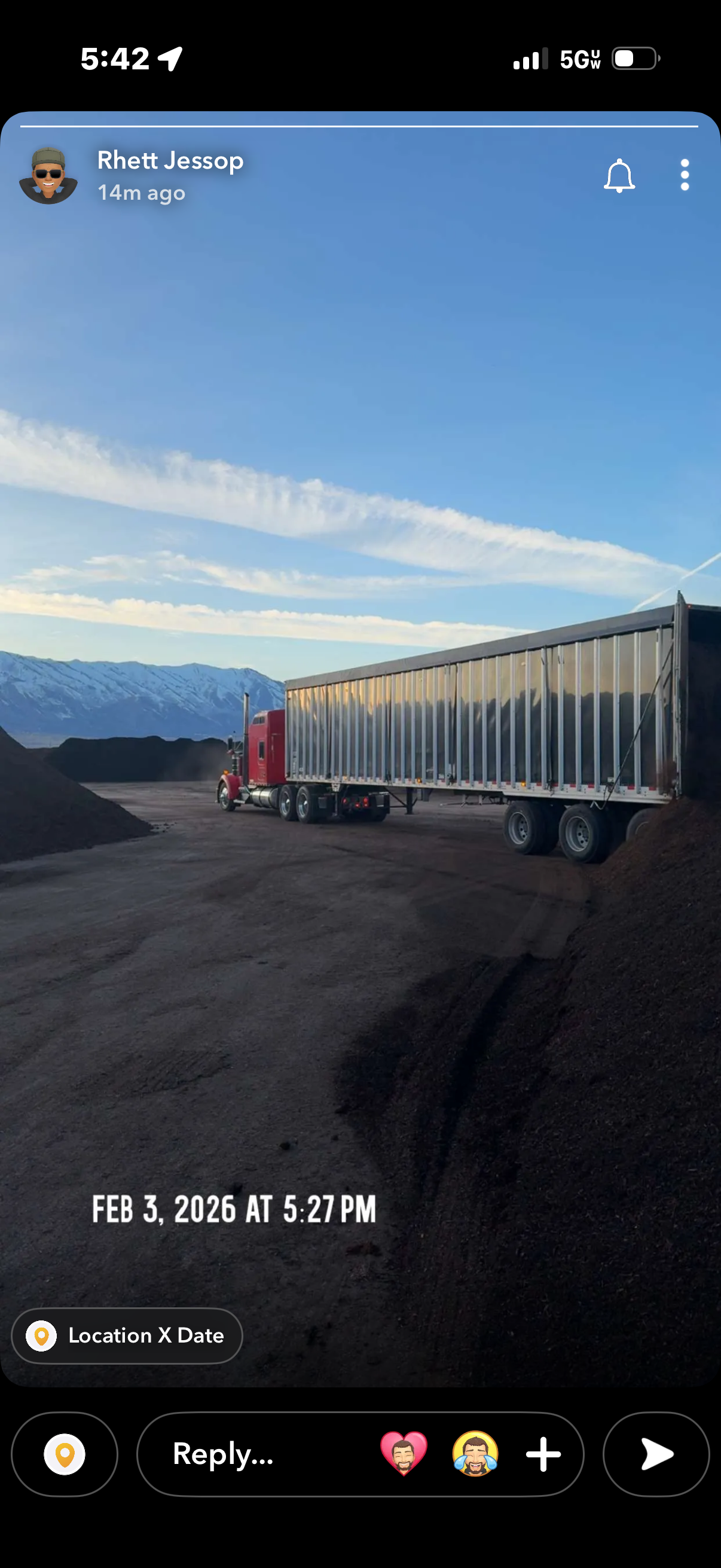 A red semi-truck with a silver trailer parked on dirt, mountains in the background, and a partly cloudy sky.