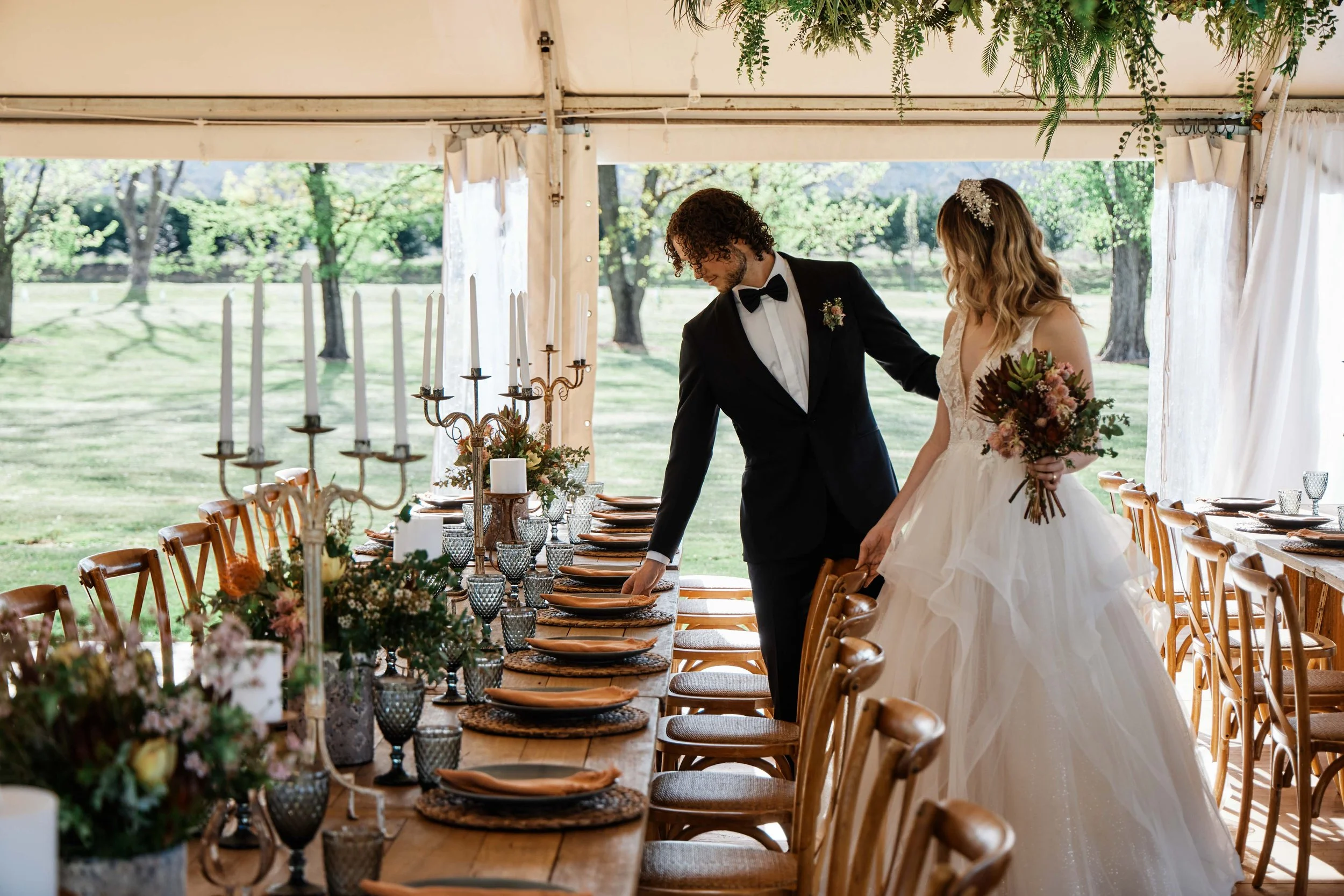 A bride and groom setting up a wedding reception table outdoors under a white tent, with trees in the background. The bride is holding a bouquet of flowers and wearing a white wedding dress. The groom is in a black tuxedo. The table is decorated with candles, flowers, and glassware.