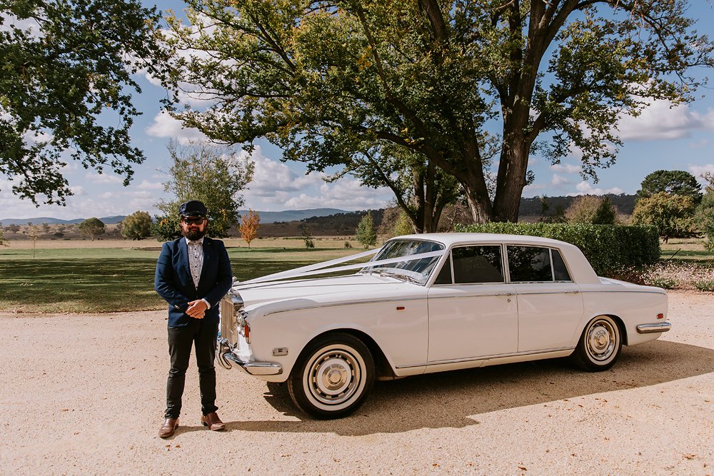 A man dressed in a suit and sunglasses standing beside a white vintage luxury car, outdoors with a tree and open landscape in the background.