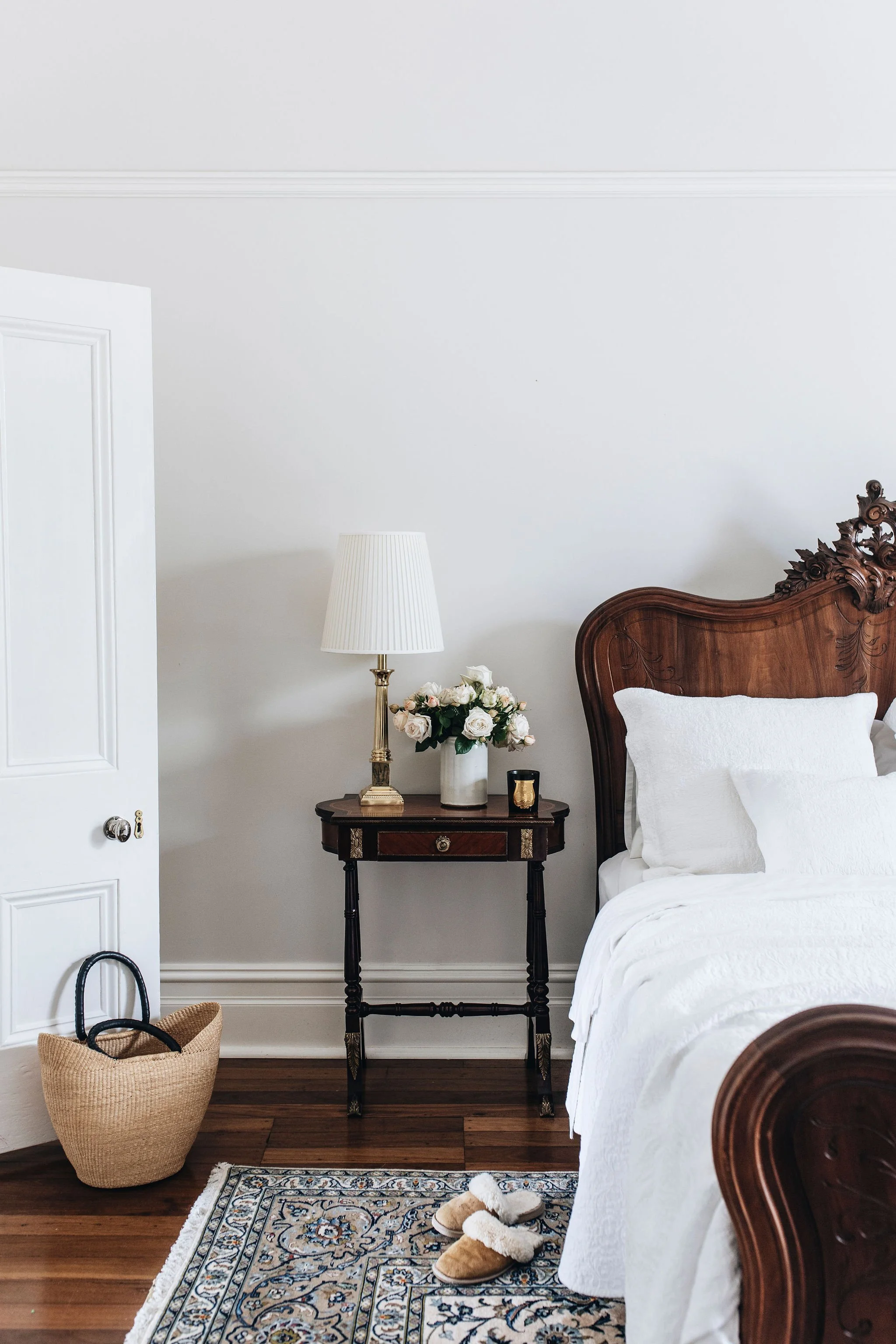 A bedroom with a wooden bed frame with white bedding, a bedside table with a floral lamp, a vase of white roses, a small candle, a basket on the floor, a pair of slippers, and a patterned rug.