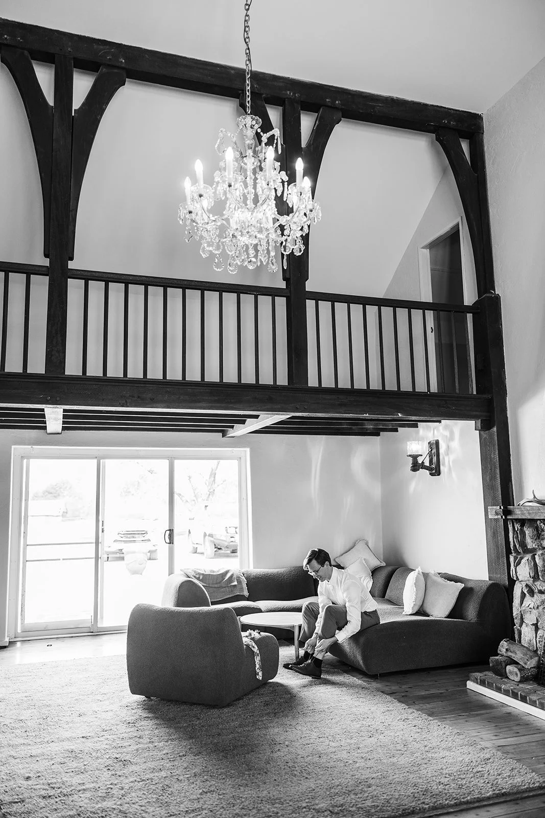 A man sits on a sofa in a living room, tying his shoes. The room has large glass sliding doors, a chandelier hanging from the ceiling, and a stone fireplace on the right. There are pillows on the sofa and a chair nearby, with a carpet on the floor.