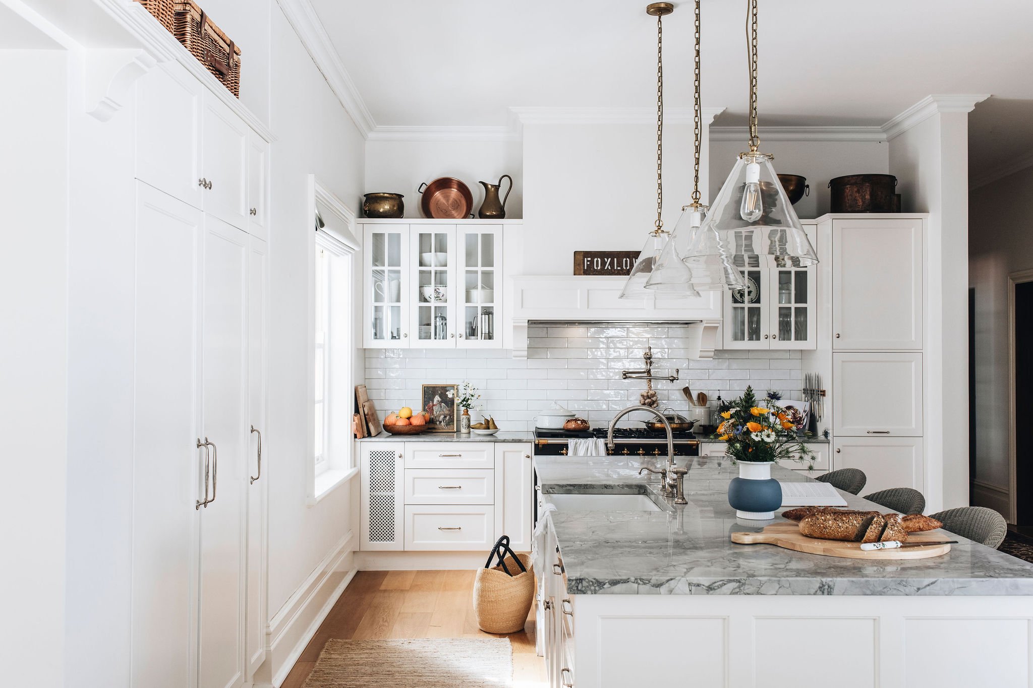 A bright white kitchen with white cabinets, a marble countertop, and a window. Decor includes copper pots, a bouquet of flowers, a cutting board with bread, and a kitchen faucet over the sink.