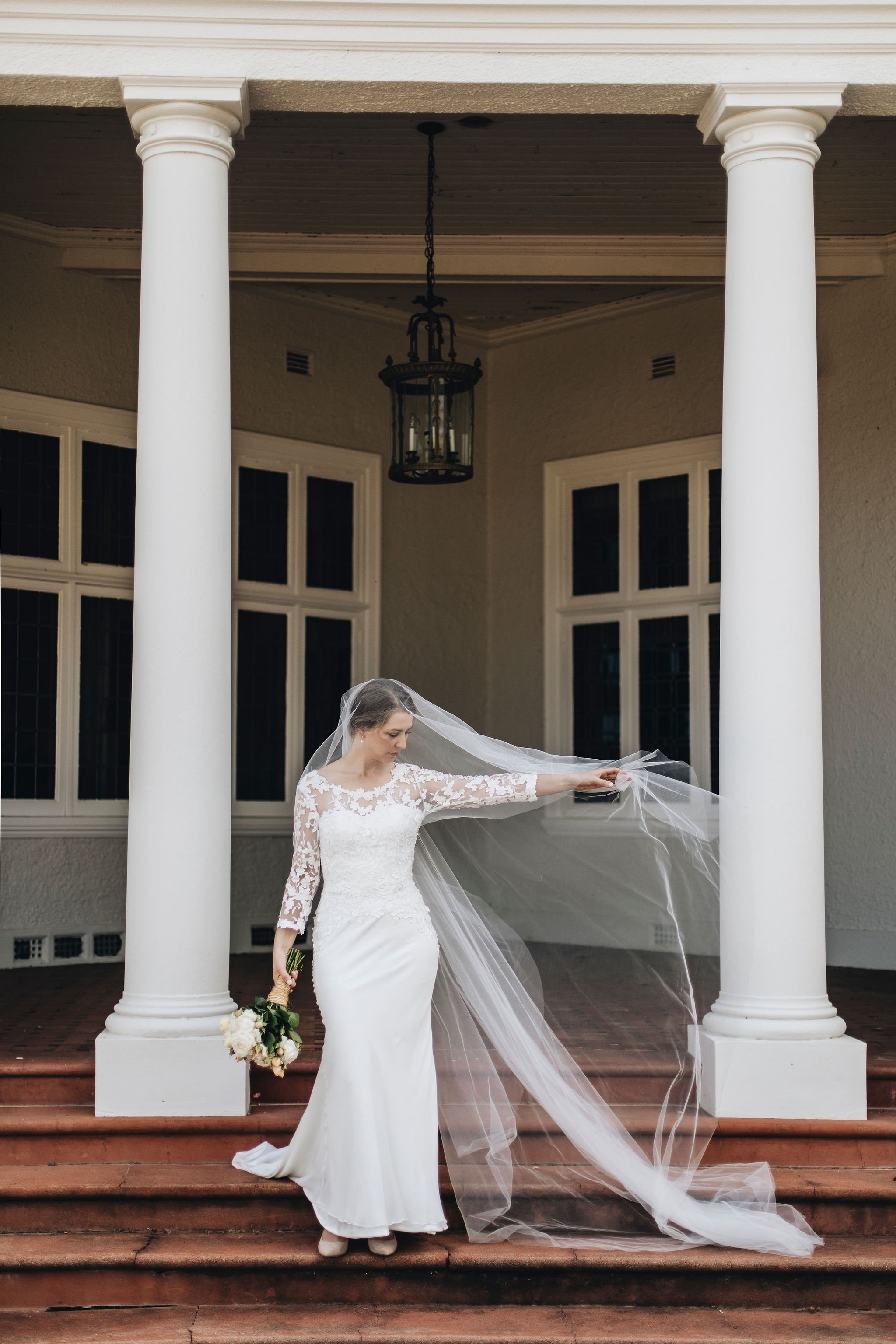 Bride in a white wedding dress holding a bouquet and adjusting her veil on the steps of a building with large white columns.