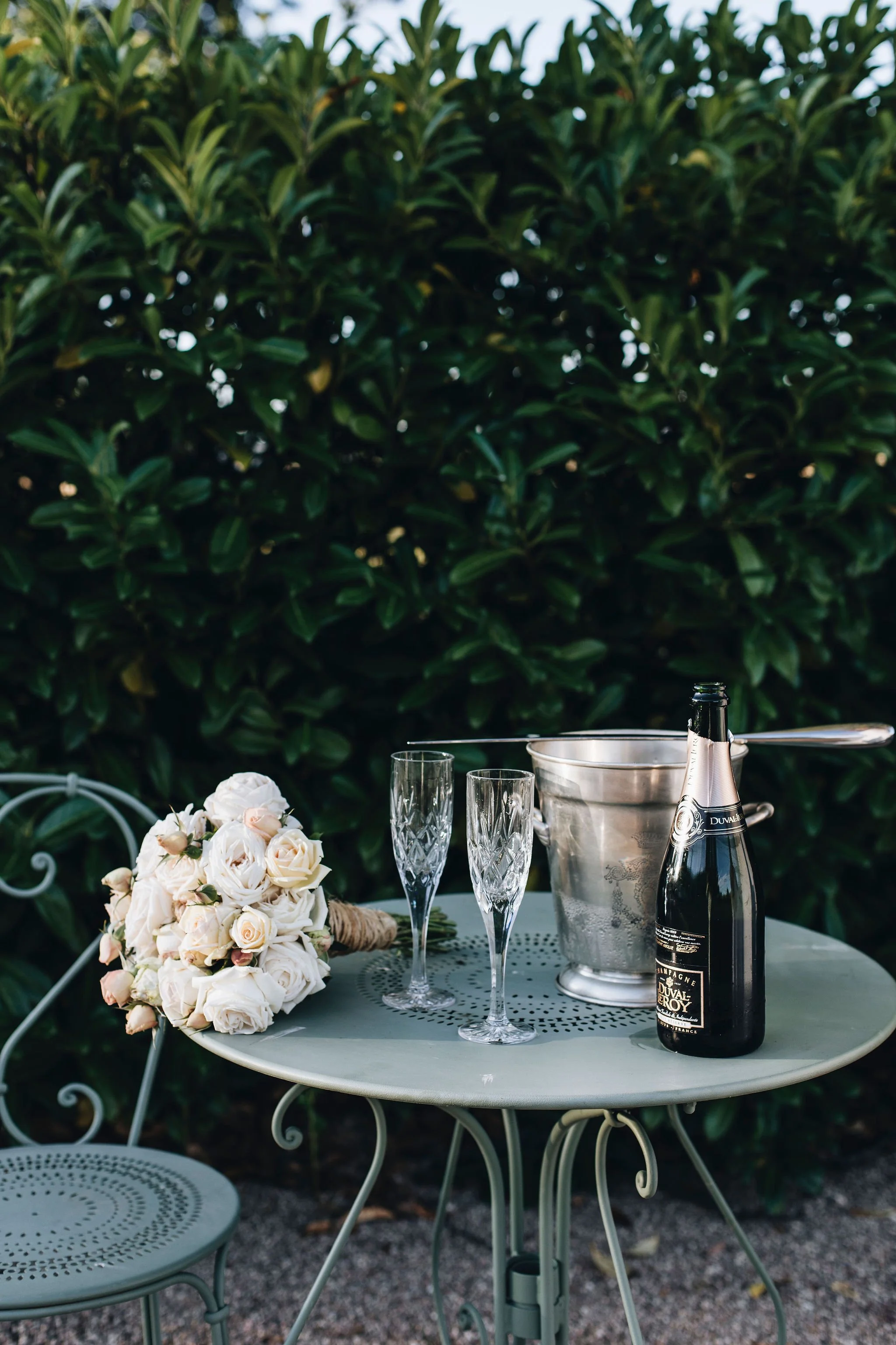 A small metal table with a matching metal chair, placed outdoors near a green leafy hedge. On the table, there is a bouquet of light-colored roses and ranunculus, two empty crystal champagne glasses, a silver ice bucket with a pair of tongs, and a bottle of sparkling wine.