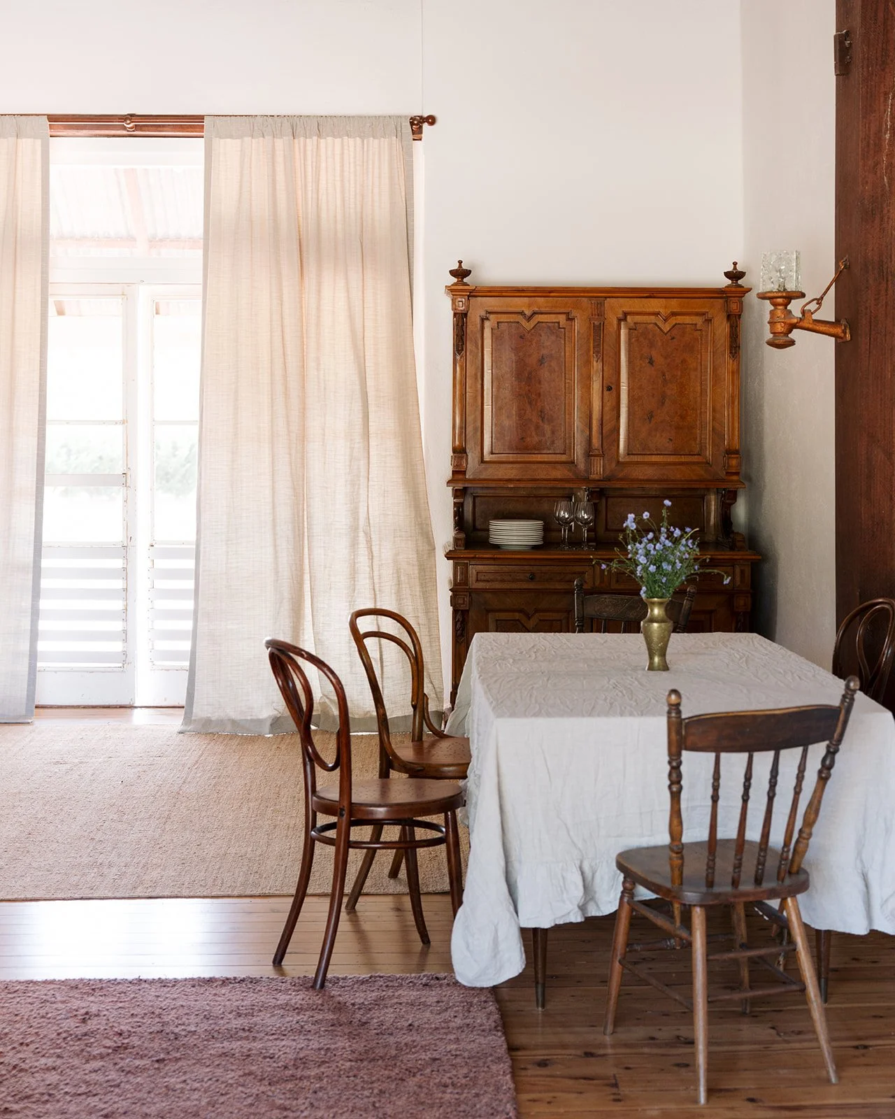 Dining room with a wooden table covered with a white tablecloth, four wooden chairs, a wooden cabinet in the background, a vase with flowers on the table, and a sliding door with curtains and a screen door outside.