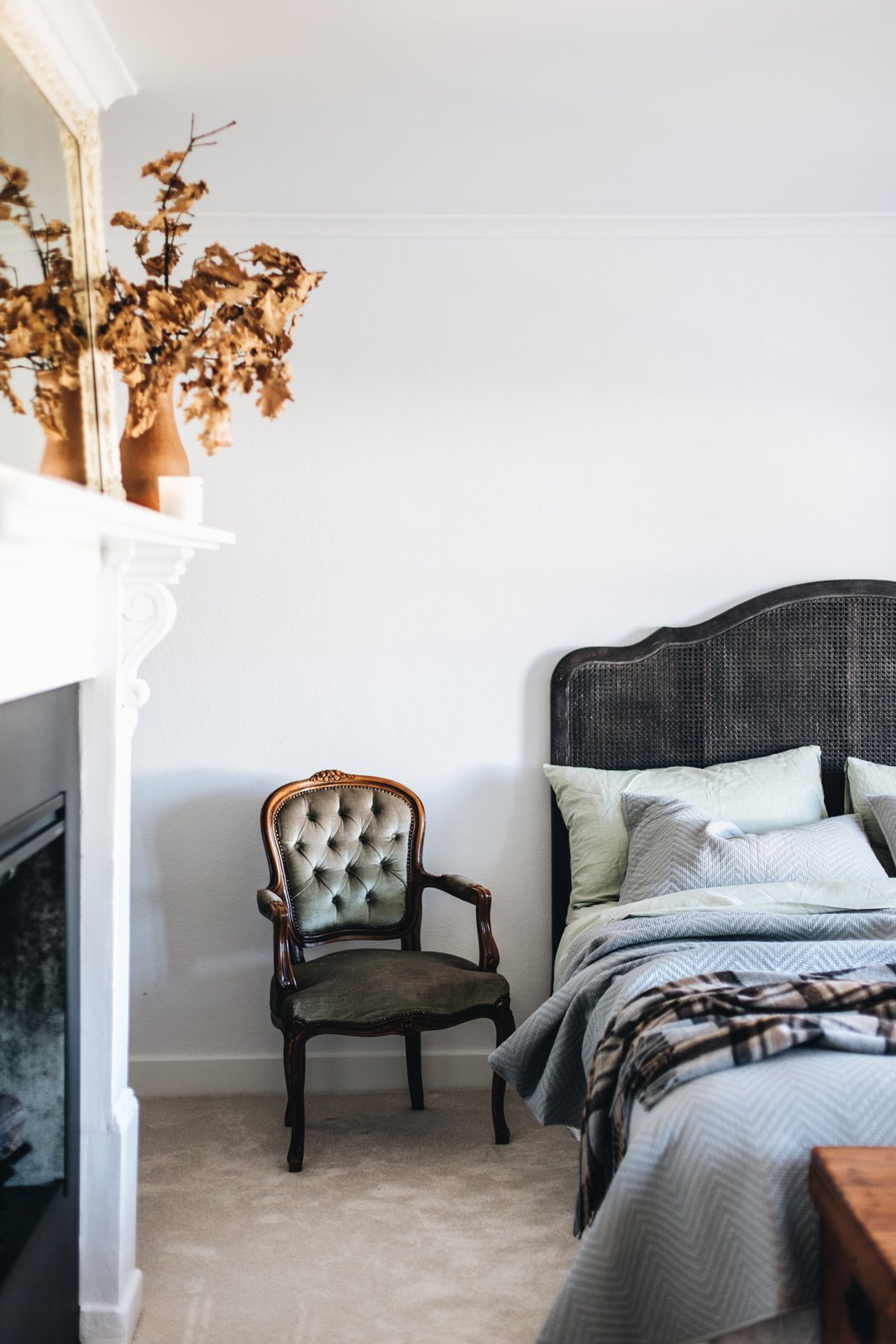 A bedroom corner with a bed, black headboard, and multiple pillows; an antique upholstered armchair with carved wood frame; a white wall, a white fireplace mantle with dried branches and a white candle; beige carpeted floor.