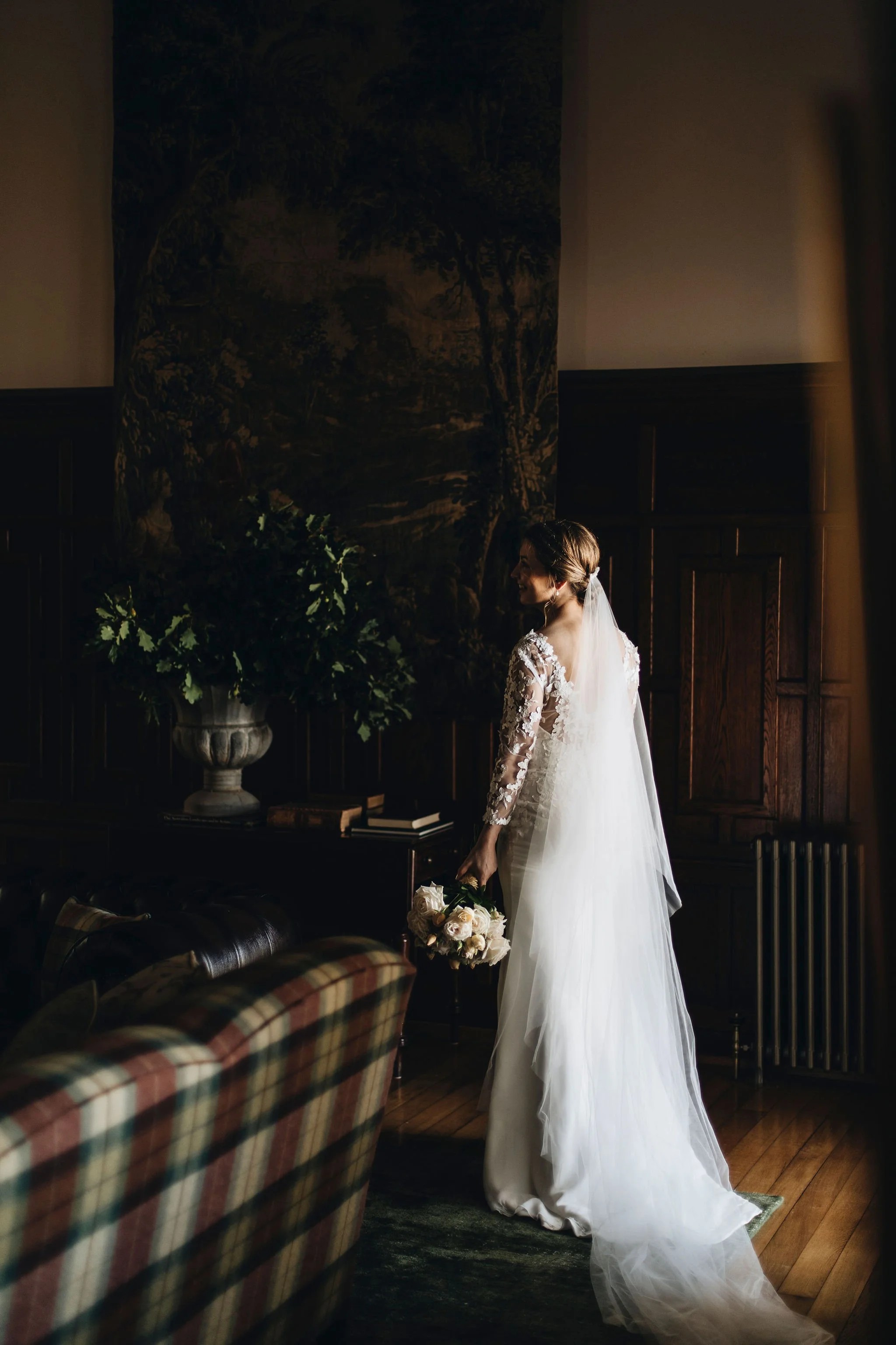 A bride in a white wedding gown holding a bouquet of roses, standing in a dimly lit room with wooden paneling and a large painting or mural on the wall.