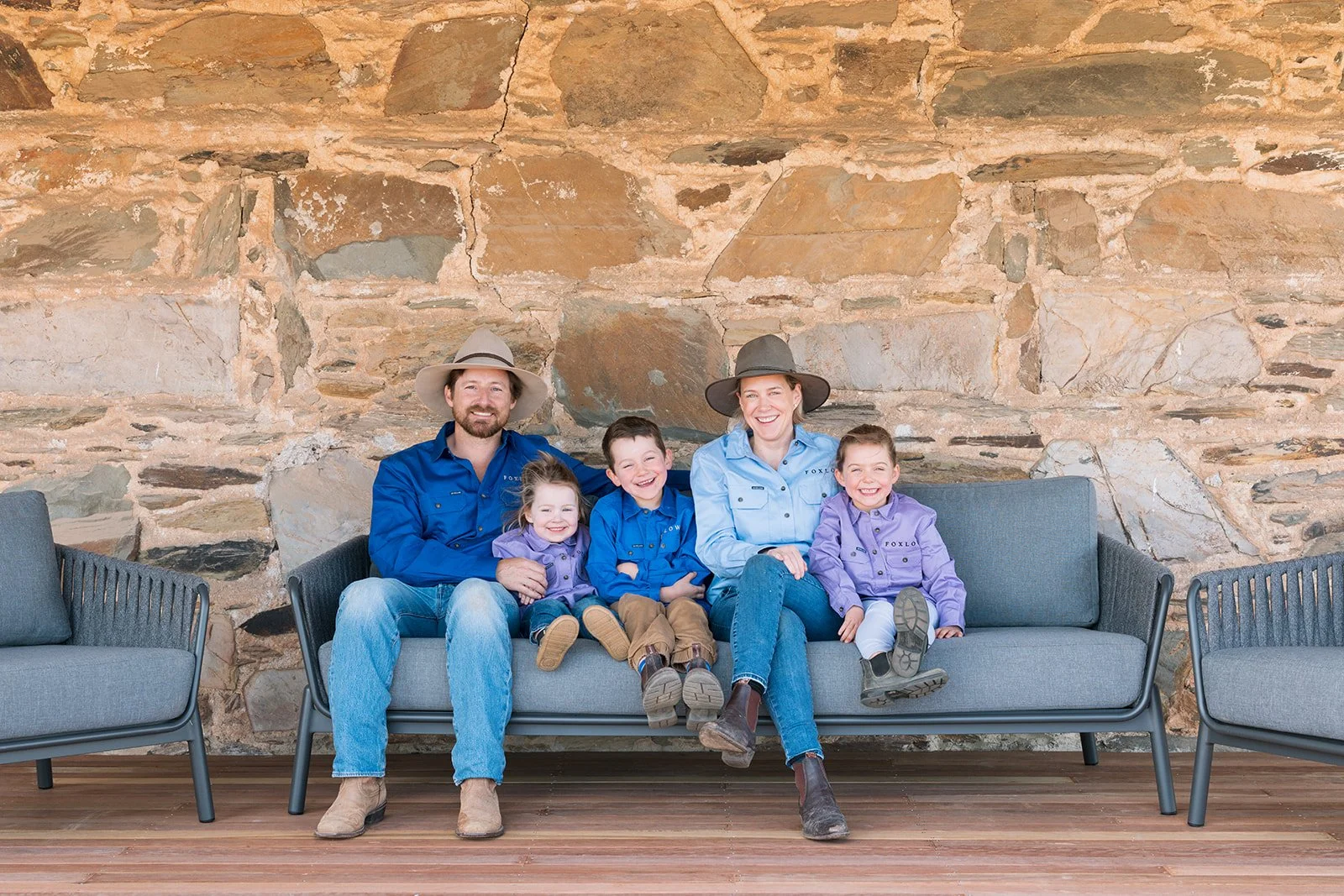 A family of six sitting on a gray outdoor sofa against a stone wall. The family includes two adults and four children, all smiling and dressed in blue and purple shirts, with the children wearing shirts labeled 'TOXLO'. The adults are wearing wide-brimmed hats.